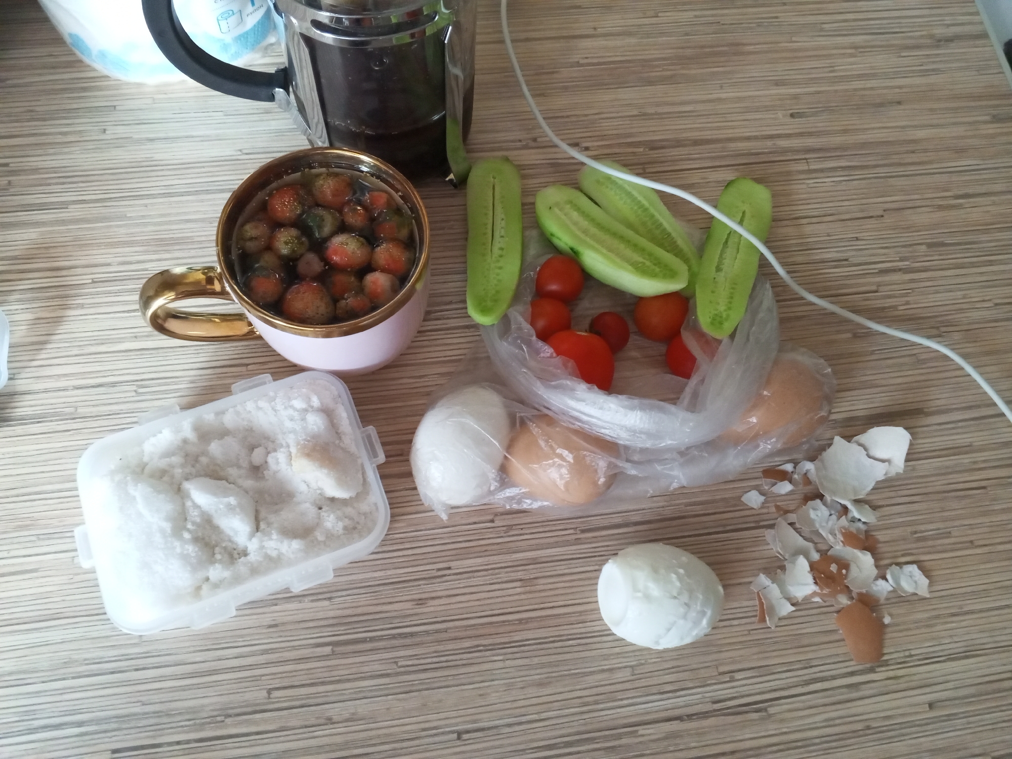On a wooden table, a collection of food items and related objects are arranged. In the upper left is a stainless steel French press coffee maker. To its right, a light pink mug with a gold rim holds small, red fruits, possibly berries or small tomatoes, submerged in liquid. In front of the mug is a white plastic container filled with a white granular substance, likely salt or sugar. To the right of the mug, there are sliced cucumbers and cherry tomatoes, some of which are partially enclosed in a clear plastic bag. Also in the bag are two whole eggs, one light brown and one white. To the right of the bag, there are several pieces of cracked eggshells. Below the bag, a peeled hard-boiled egg sits on the table, and a single whole hard-boiled egg is also visible to the left of the eggshells. A white electrical cord snakes across the scene. The image was captured on a table in Chistopol, Russia.