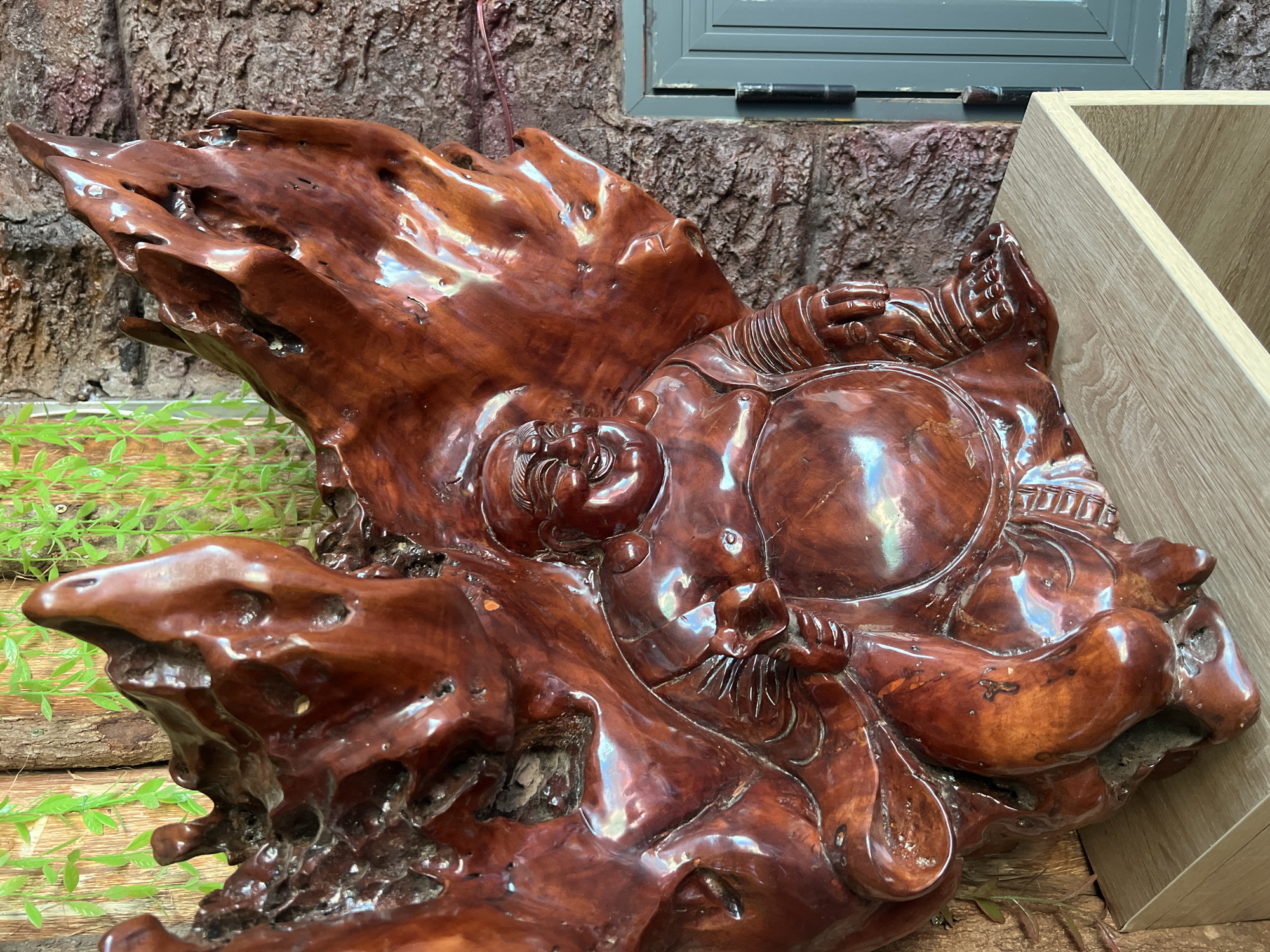 A highly polished, dark reddish-brown wooden carving dominates the frame. The carving appears to be in the form of a reclining figure, with suggestions of a face, round belly, and limbs. The wood has many natural imperfections and contours that have been incorporated into the piece. The carving is situated on what seems to be a wooden surface, partially obscured by small green plants. The background includes a textured wall, possibly of artificial rock, and a grey window frame. A light-colored wooden box is also positioned to the right of the carving. There is no visible evidence of a city, Bac Natural Heritage Area, or Trang, Vietnam in the image.