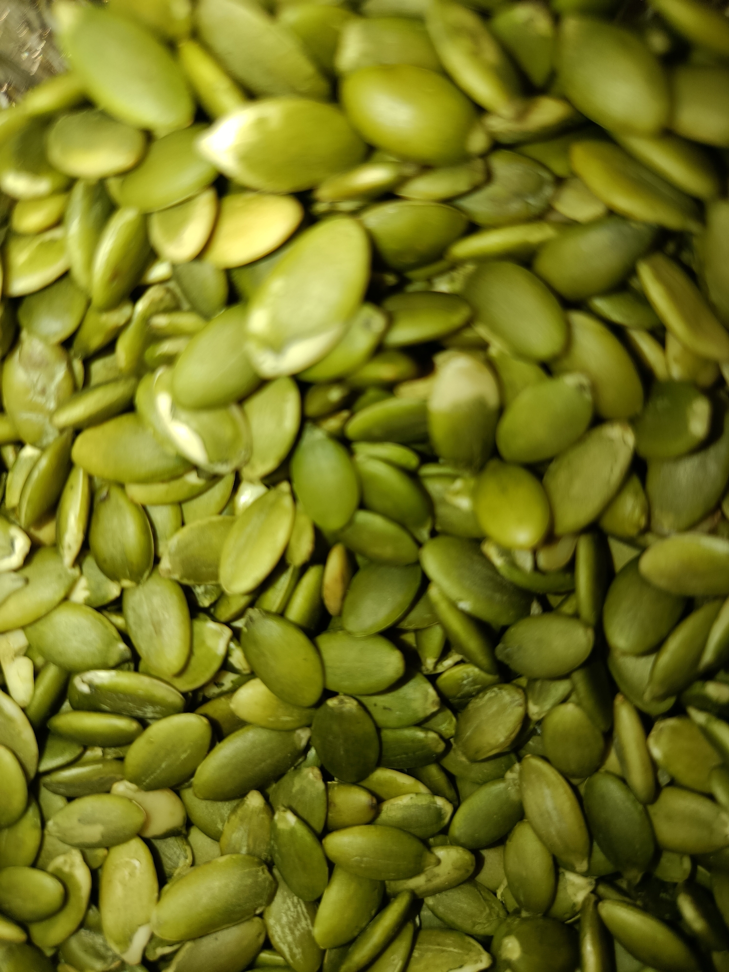The image is a close-up, top-down view of a large pile of raw, shelled pumpkin seeds. The seeds are oval-shaped and a vibrant green color, with some showing lighter creamy-white interiors where they have split or are viewed from the side. They are densely packed and overlap each other, creating a textured surface. The lighting is somewhat harsh, causing highlights and shadows that give depth to the pile. The focus is sharp on the seeds in the foreground, gradually softening towards the background, suggesting a shallow depth of field. There is no identifiable background or context provided to link this image to Tashkent, Uzbekistan, or any specific urban environment.