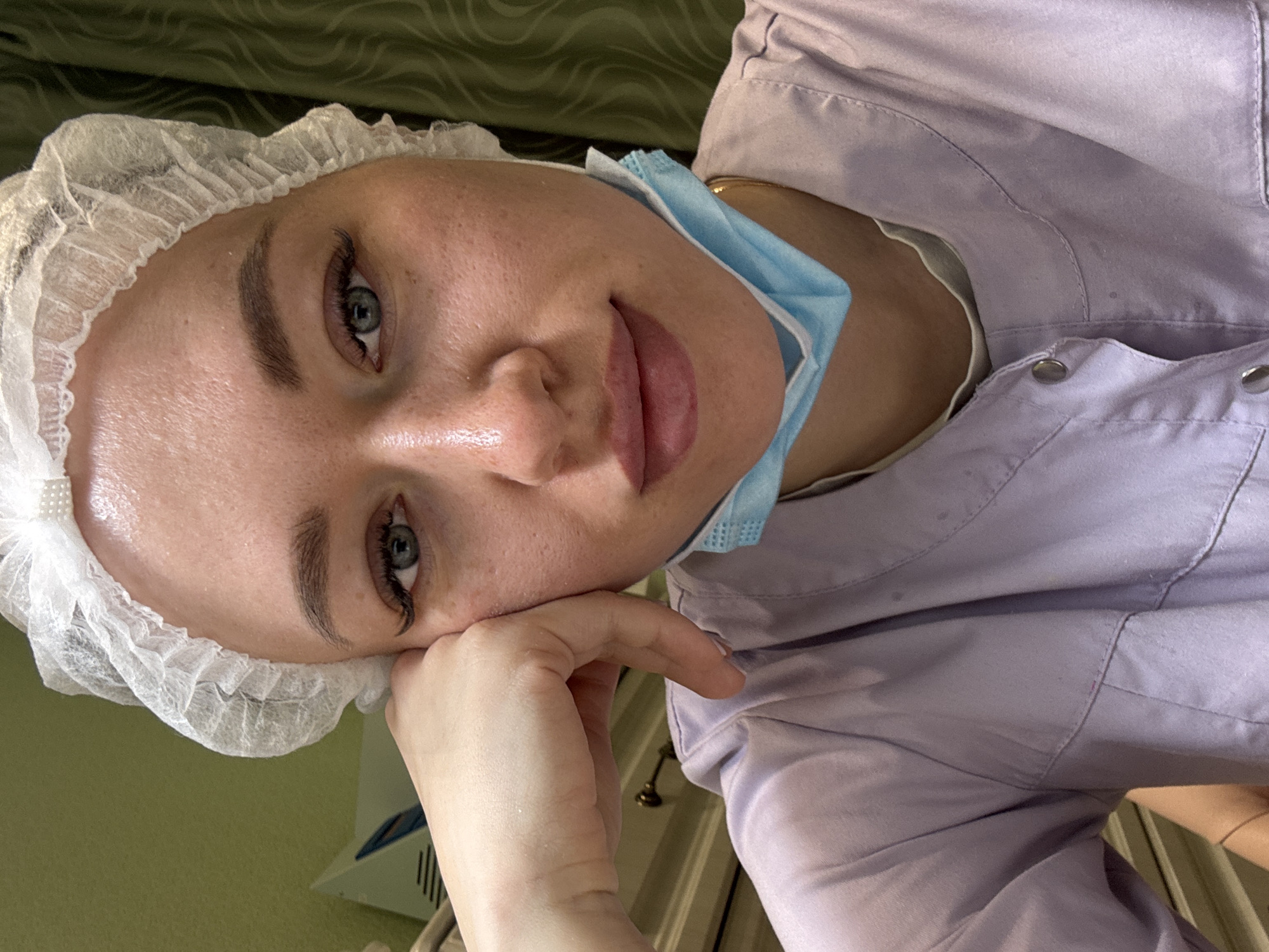 A woman wearing a white surgical cap and a blue surgical mask is visible. The mask is pulled down, revealing her lips. She's wearing a light purple buttoned shirt or jacket. Her left hand is near her face, seemingly resting it. The background shows a green wall. The location is in the city of Ферма, Russia.