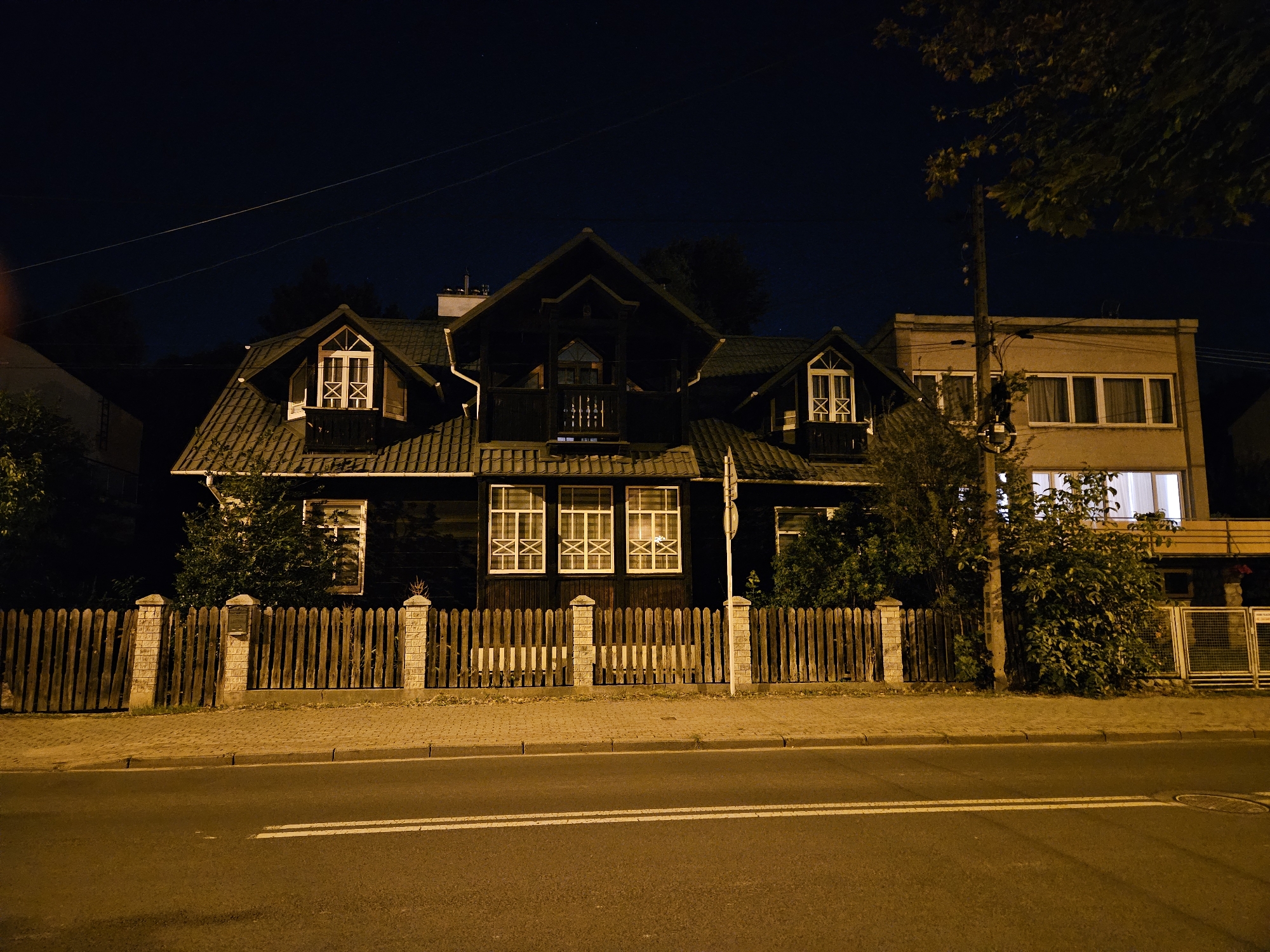 The image shows a two-story wooden house with a dark exterior, typical of a traditional Polish architectural style, at night. The house features multiple windows, some with decorative cross-shaped elements. A wooden fence with stone pillars separates the property from the street. Adjacent to the wooden house is a lighter-colored, more modern building with larger windows. The street in front of the houses is paved and has white lane markings. The sky is dark, indicating nighttime. The scene is illuminated by artificial lights, casting a warm glow on the buildings and pavement. The overall impression is a quiet, residential street at night, with a mix of older and newer architecture. The specific location of this house in Rzeszów, Poland cannot be determined solely from the visual information provided in the image.