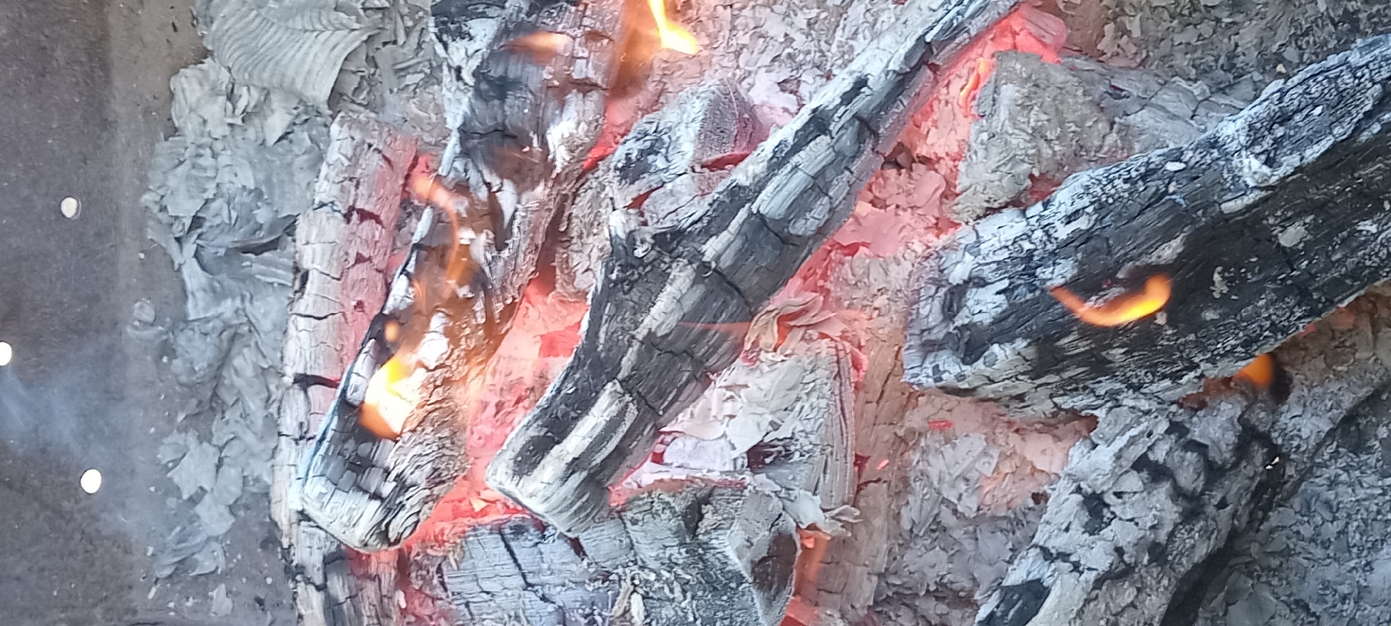 The image displays a close-up view of glowing embers and charred logs in what appears to be the aftermath of a fire. The scene is dominated by shades of gray, black, and vibrant orange-red from the hot coals. Several pieces of partially burned wood are visible, covered in ash and showing cracks from the heat. Small flames lick at the edges of some logs, indicating residual heat. The environment suggests an outdoor setting, possibly a fire pit or a collapsed structure. There are no people or animals present in the image. The specific location in Koviahy, Ukraine, is not visually discernible from the image itself.