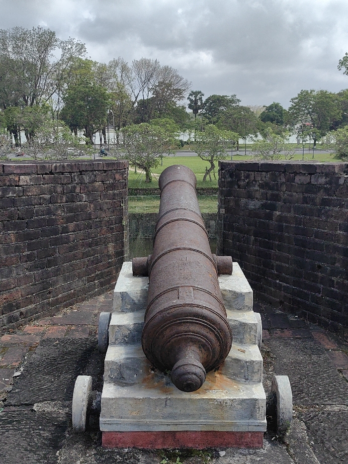 A rusty, dark brown cannon is positioned centrally, pointing forward, resting on a multi-tiered grey stone carriage with two visible grey wheels. It is flanked by two dark red-brown brick walls, which show signs of age. The immediate ground is paved with dark, weathered stones, and a red base is visible beneath the cannon's carriage. Beyond the brick walls, a narrow grassy area and a water-filled moat or trench separate the foreground from a paved road. The road features white markings and a parallel white barrier. In the mid-distance on the road, small figures on what appear to be motorbikes or bicycles are discernible, including a distinct dark blue-green scooter. The background is dominated by a dense line of trees with varying foliage, some green and others bare, with a large body of water visible behind them. The sky is uniformly overcast and grey. This scene is located in Phú Xuân Ward, Vietnam.