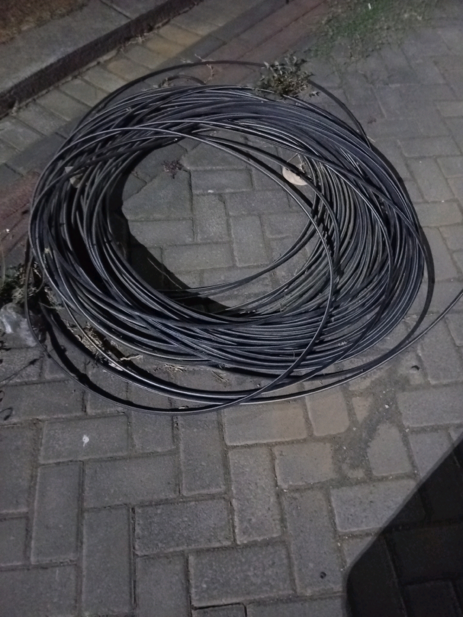 A coiled black cable lies on a paved surface composed of interlocking grey bricks. The cable appears to be a heavy-duty electrical or communication cable, tightly wound into a circular shape, filling a significant portion of the image. The surrounding environment is dimly lit, suggesting it might be nighttime or dusk. In the background, to the left and top, there are hints of vegetation and possibly a concrete structure, which could be a pathway or the edge of a building. The paved area shows typical wear and tear of an outdoor urban setting. The location is specified as the city of Malang, Indonesia.