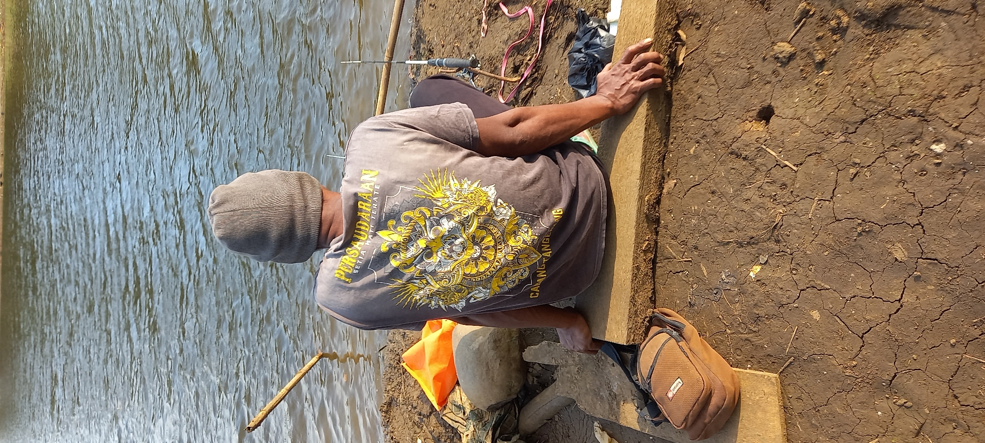 A person wearing a grey beanie and a grey t-shirt with yellow graphic text and a design on the back is sitting on a wooden plank by the water. The text on the shirt reads "PERSAUDARAAN SETIA HATI TERATE". A brown satchel is placed on the ground to the left of the person. The background consists of water and a muddy, cracked ground with some rocks and debris. A fishing rod is visible in the water behind the person. The scene is set outdoors during daylight.