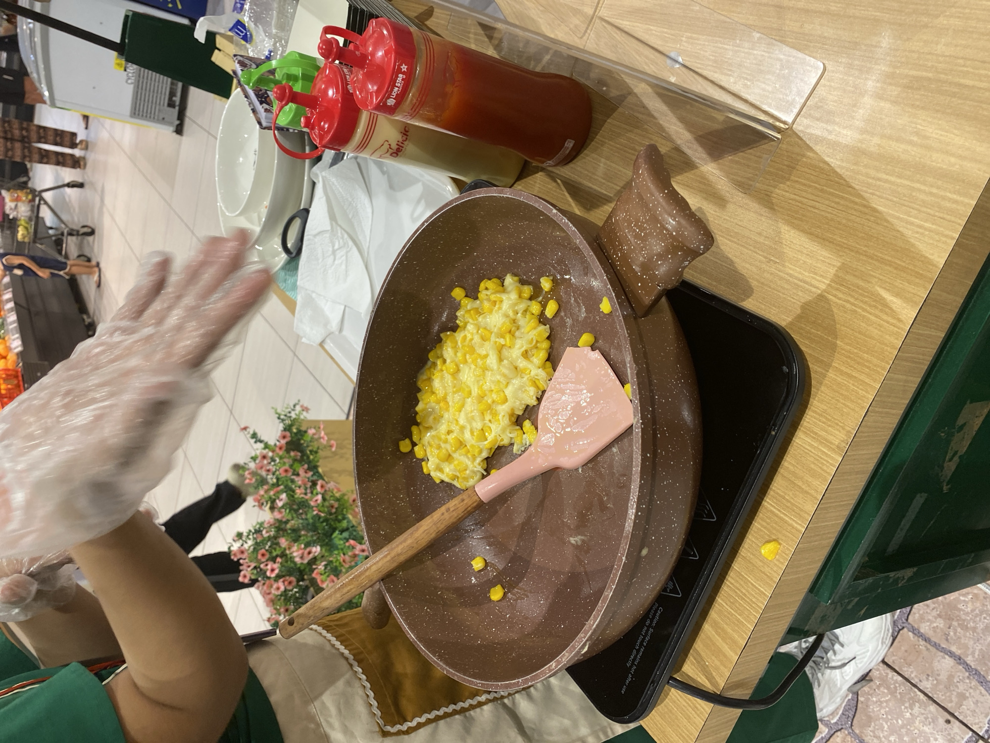 In East Jakarta, Indonesia, a person wearing a green shirt and an apron is cooking scrambled corn in a brown pan on an induction cooktop. They are wearing clear plastic gloves and are holding a pink spatula. Next to the pan are two red squeeze bottles, one with a yellow label and the other with a red label, and a white bowl and plate. In the background, a shopping cart and other shoppers are visible in a supermarket setting. There are decorative pink flowers in a planter behind the person.