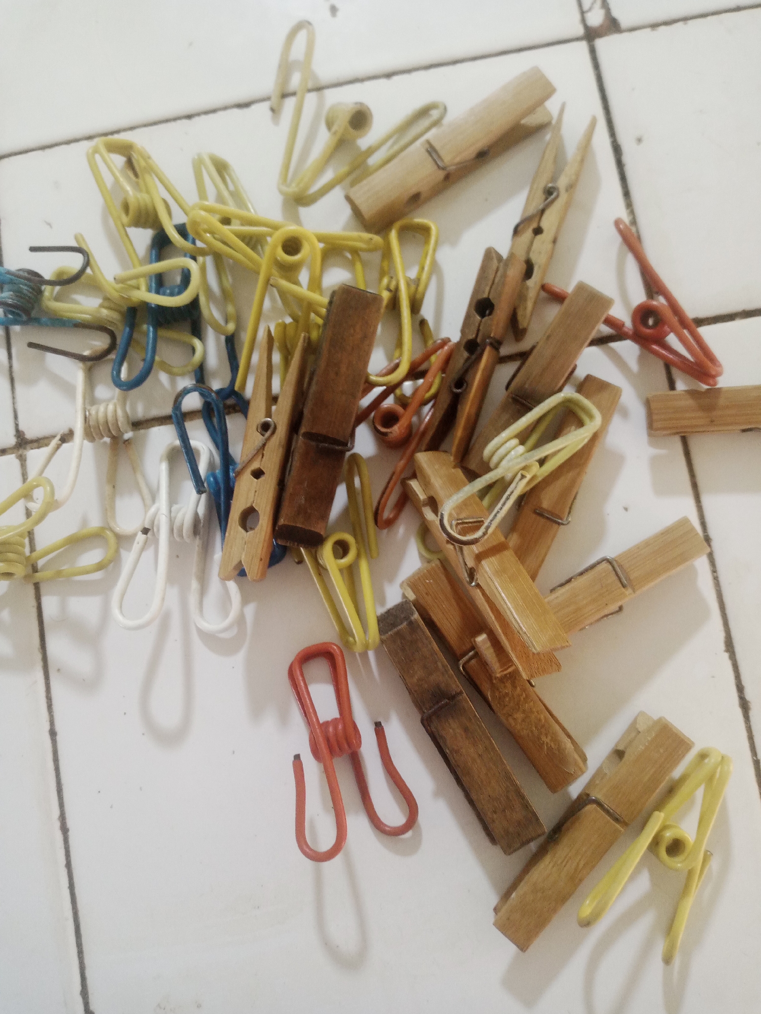 The image shows a collection of clothespins and clips scattered on a tiled surface, possibly a floor or countertop, in what appears to be a residential setting. The objects are of various colors, including natural wood, yellow, blue, white, and red. Some of the clothespins are made of wood, while others are metal or plastic. They are arranged in a disorganized manner, suggesting they have been used and placed down casually. The tiled surface has grout lines and appears to be somewhat worn, with some minor debris visible. The lighting is artificial and casts shadows on the objects. There are no people visible in the image. This scene does not provide any specific indicators of its location within the city of La Tagua, Venezuela.