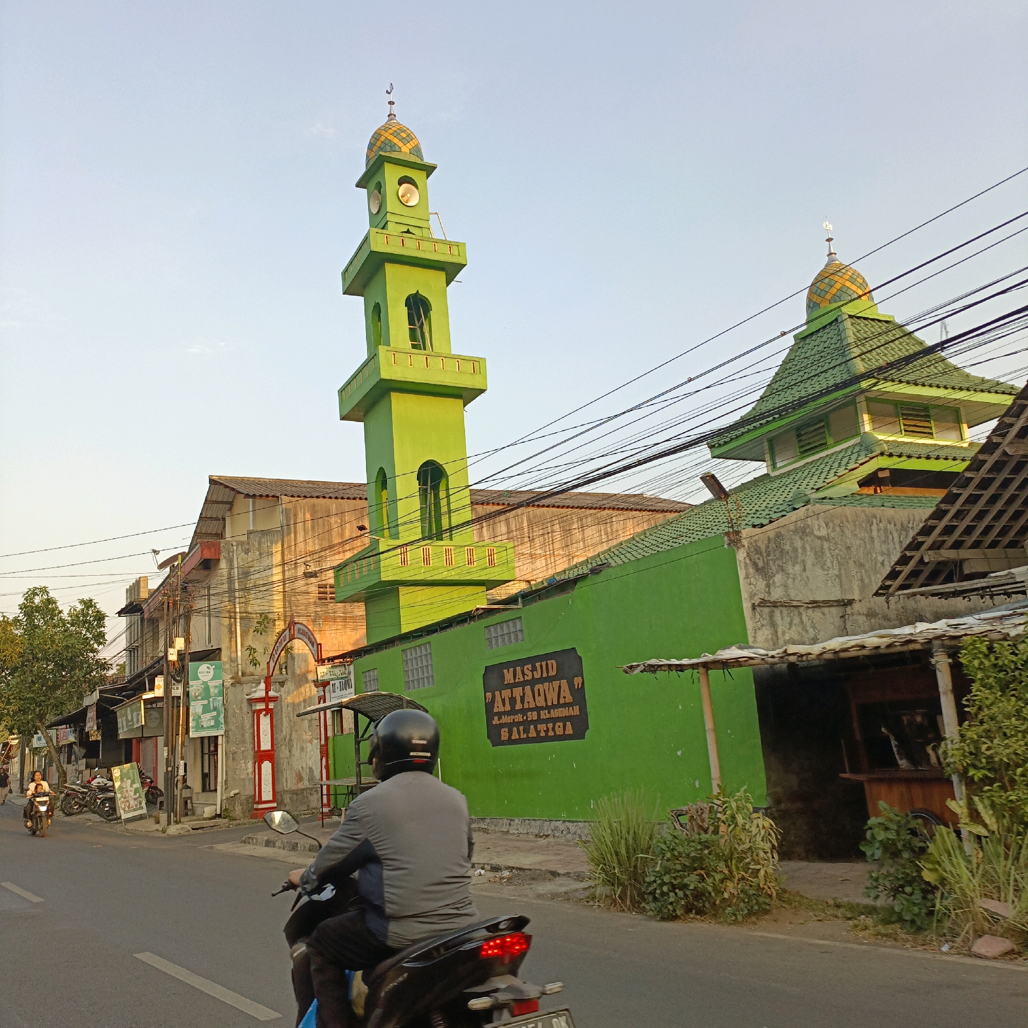 The image captures a street scene in Salatiga, Indonesia, featuring a mosque and its surroundings. The mosque, painted a vibrant green, has a prominent minaret topped with a gold and green patterned dome. To the right, another part of the mosque features a similarly designed green roof. A sign on the green wall of the mosque reads "MASJID 'ATT'AQWA" and below it, "JL. Merak No. 59 Klaseman Salatiga".
In the foreground, two individuals are riding on a motorcycle, viewed from behind. The rider wears a black helmet and a grey jacket, while the passenger appears to be wearing dark clothing. They are traveling on an asphalt road.
The street is lined with buildings, some of which appear to be shops or small businesses, with awnings and signage visible. A few other motorcycles are parked or in motion on the street. Trees and vegetation are present along the roadside, and utility wires crisscross the sky. The lighting suggests it is late afternoon or early evening, with the sun casting a warm glow on some of the buildings.