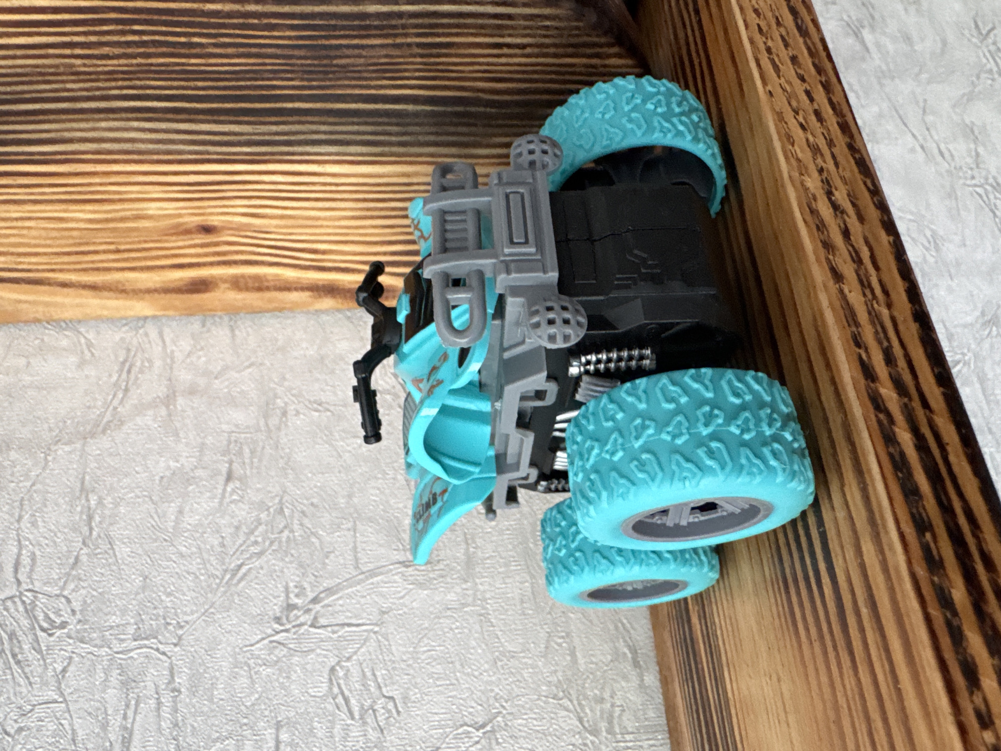 A teal and gray toy quad bike is positioned on a wooden shelf against a textured white wall and a vertical wooden plank. The quad bike features large teal tires with aggressive treads, a gray front bumper, and teal bodywork with decorative graphics. The background includes a white wall with a rough, crinkled texture and a vertical wooden plank with distinct grain patterns, possibly part of a wall or furniture. The scene is lit from the front, casting subtle shadows. There are no people present, and no indication of the specific location within Яромінскі сельскі Савет, Belarus.