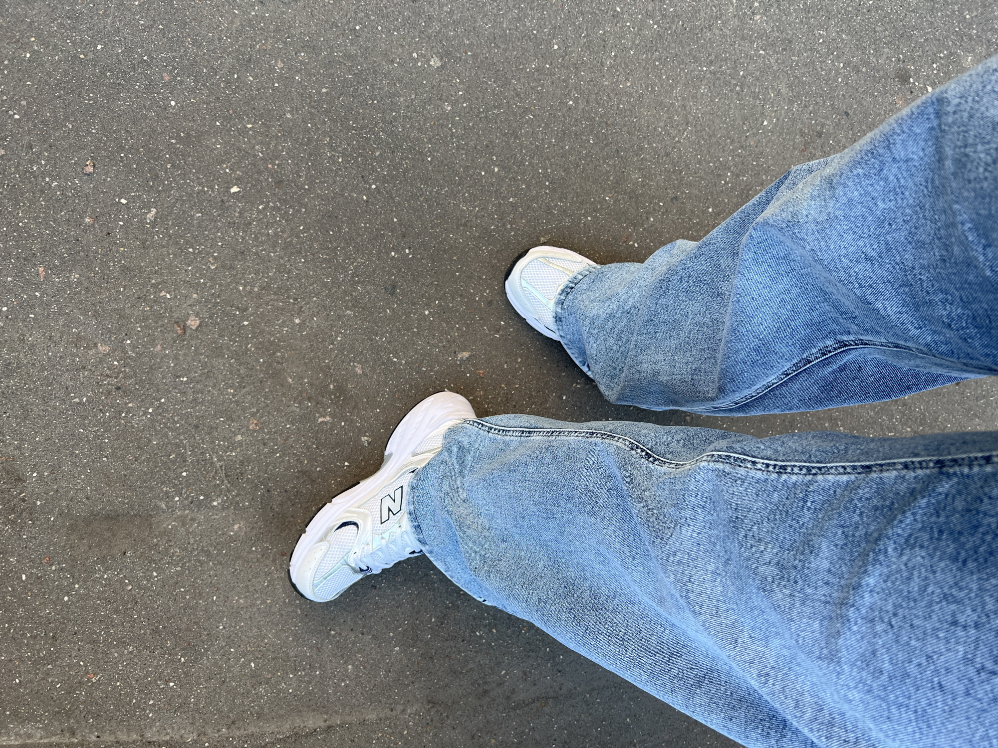 An overhead shot shows a person's legs from the knees down, clad in light blue, wide-leg denim jeans and white athletic shoes with dark accents. The person is standing on a dark gray asphalt surface speckled with small white and reddish-brown stones. The lighting suggests an outdoor environment. The location is identified as Khimki, Russia.