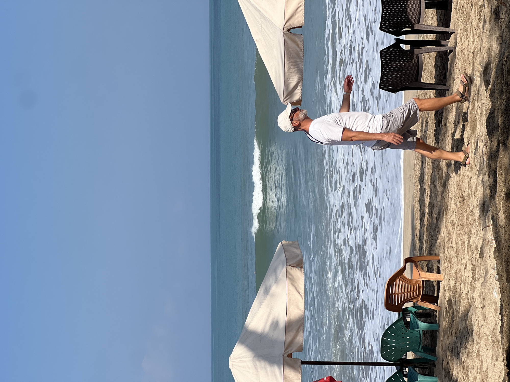 A man walks along a sandy beach in Kuta, Indonesia, with the ocean visible in the background. He is wearing a white t-shirt, grey shorts, a white baseball cap, and sandals. The man is walking from right to left in the frame. Two large umbrellas provide shade on the beach, and several beach chairs are scattered on the sand. The ocean has moderate waves. The sky is clear and blue.