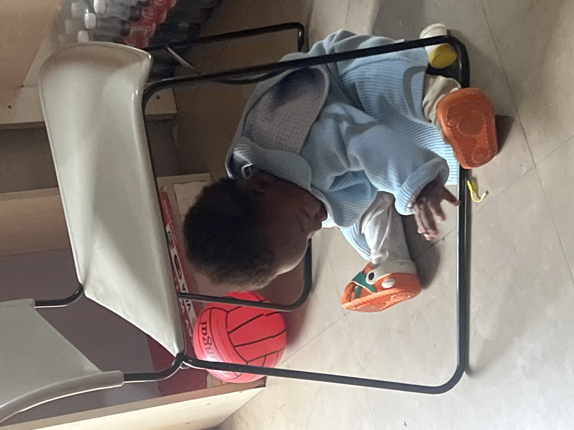 A child in a light blue hooded jacket and white pants sits in a metal-framed chair with a white seat and back. The child wears orange and white shoes and has dark hair. A red ball with black markings rests near the chair leg. The chair is on a light-colored tiled floor. The location is likely Gura Top, Nigeria, based on the prompt.