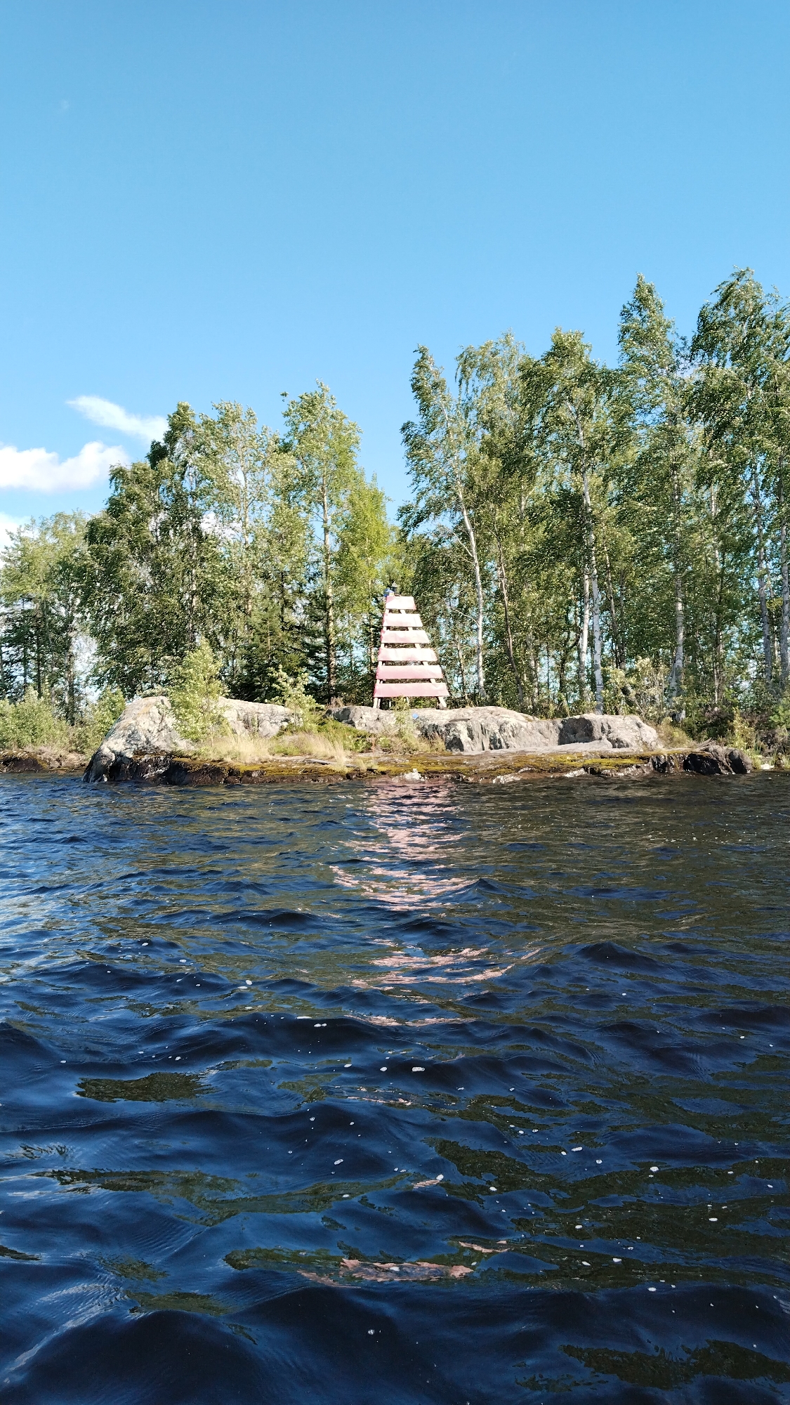 A bright, sunny day with a clear blue sky and scattered clouds is depicted. The scene is a body of water, likely a lake or river, with dark blue, choppy water in the foreground. In the background, there is a rocky island covered with green grass and a dense line of tall, green trees, primarily birch trees with white trunks. A distinctive red and white tiered structure, resembling a large ladder or a navigational marker, stands on the rocks in the middle ground, positioned among the trees. The reflection of the structure and the trees can be seen on the surface of the water. There are no people visible in the image. The overall impression is a natural, serene landscape.