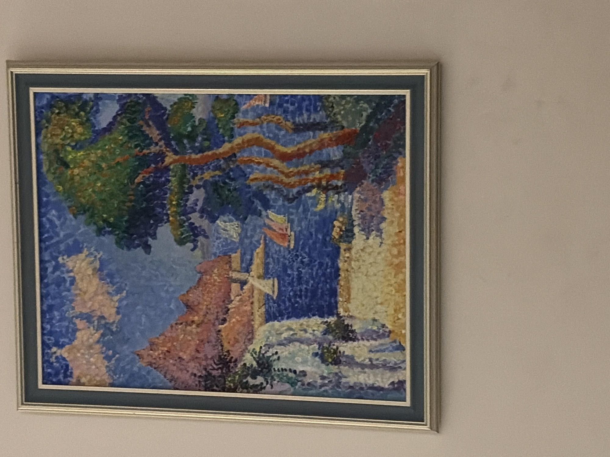 The image shows a framed painting hanging on a light-colored wall. The painting depicts a seascape with sailboats, rocky hills, a prominent tree, and a coastline. The style appears to be pointillist or post-impressionist, with visible dabs of color. The frame is silver with a dark mat. The painting is centrally located on the wall. No people or specific landmarks that would indicate a location in Germasogeia Municipality, Cyprus, are visible in the artwork itself.