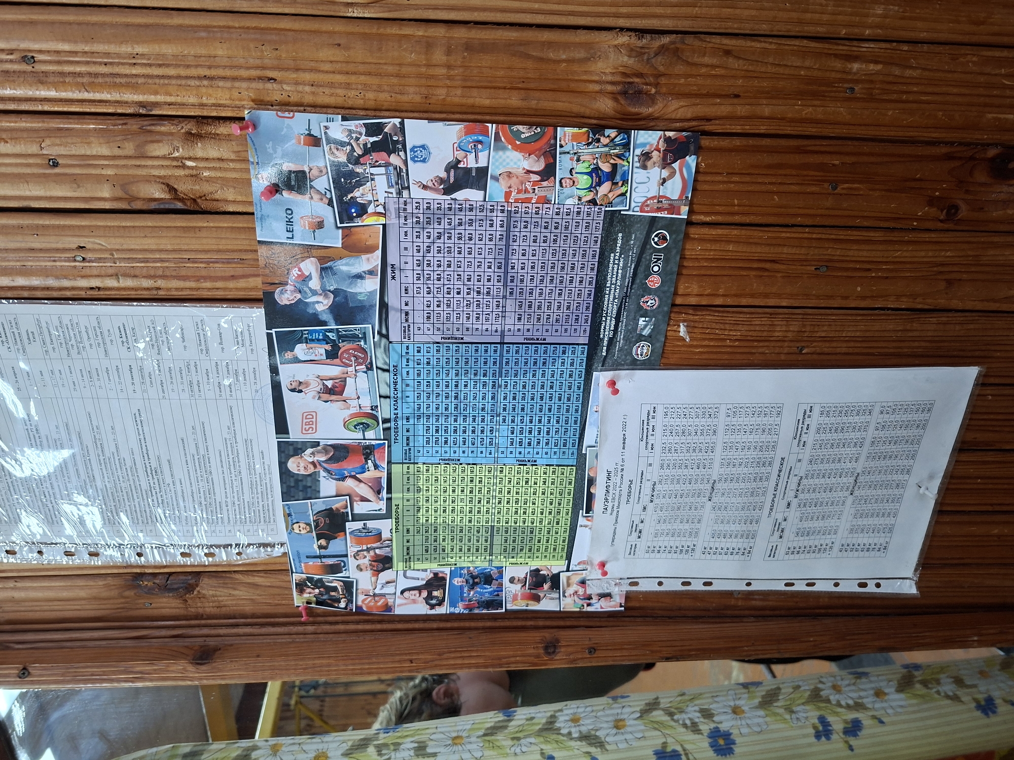 The image depicts a close-up of a wooden wall with various documents and photographs attached. The documents appear to be competition schedules and results for weightlifting, with tables containing categories like "весовые категории" (weight categories), "МСМК" (Master of Sport of Russia, International Class), "МС" (Master of Sport of Russia), "КМС" (Candidate Master of Sport), and different ranks within sporting categories. One document is titled "ПАУЭРЛИФТИНГ" and mentions "Россия" and "2022-2025 гг.". The photographs show athletes, primarily weightlifters, in action, lifting weights and posing. There are also logos of sports organizations visible. The overall impression is that of a sports club or training facility. There is no visible indication of the city of Kazan, Russia.