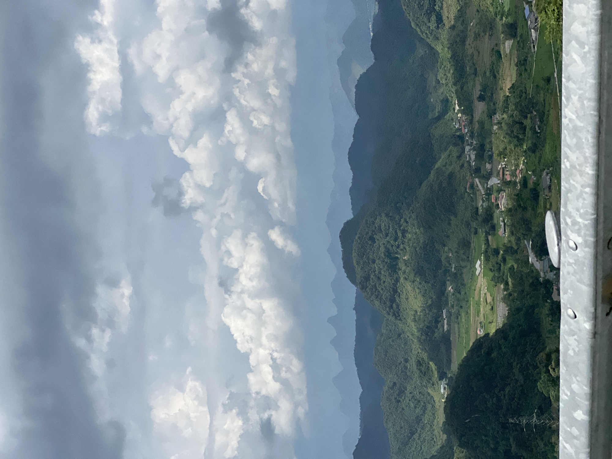 The image captures a vast landscape of rolling green mountains and valleys under a dramatic, cloud-filled sky. In the foreground, a silver metal railing with visible rivets suggests the viewpoint is from a road or overlook. The midground features densely vegetated, rounded hills, with patches of cultivated land and scattered buildings nestled within the valleys. A few distinct structures, including what appears to be a village or small settlement, are visible. The background reveals a series of progressively more distant, blue-hued mountain ranges, creating a sense of depth. The overall impression is one of a serene, rural mountainous region, likely Quản Bạ Commune in Vietnam, given the distinctive karst landscape and vegetation. No people are visible in the frame.