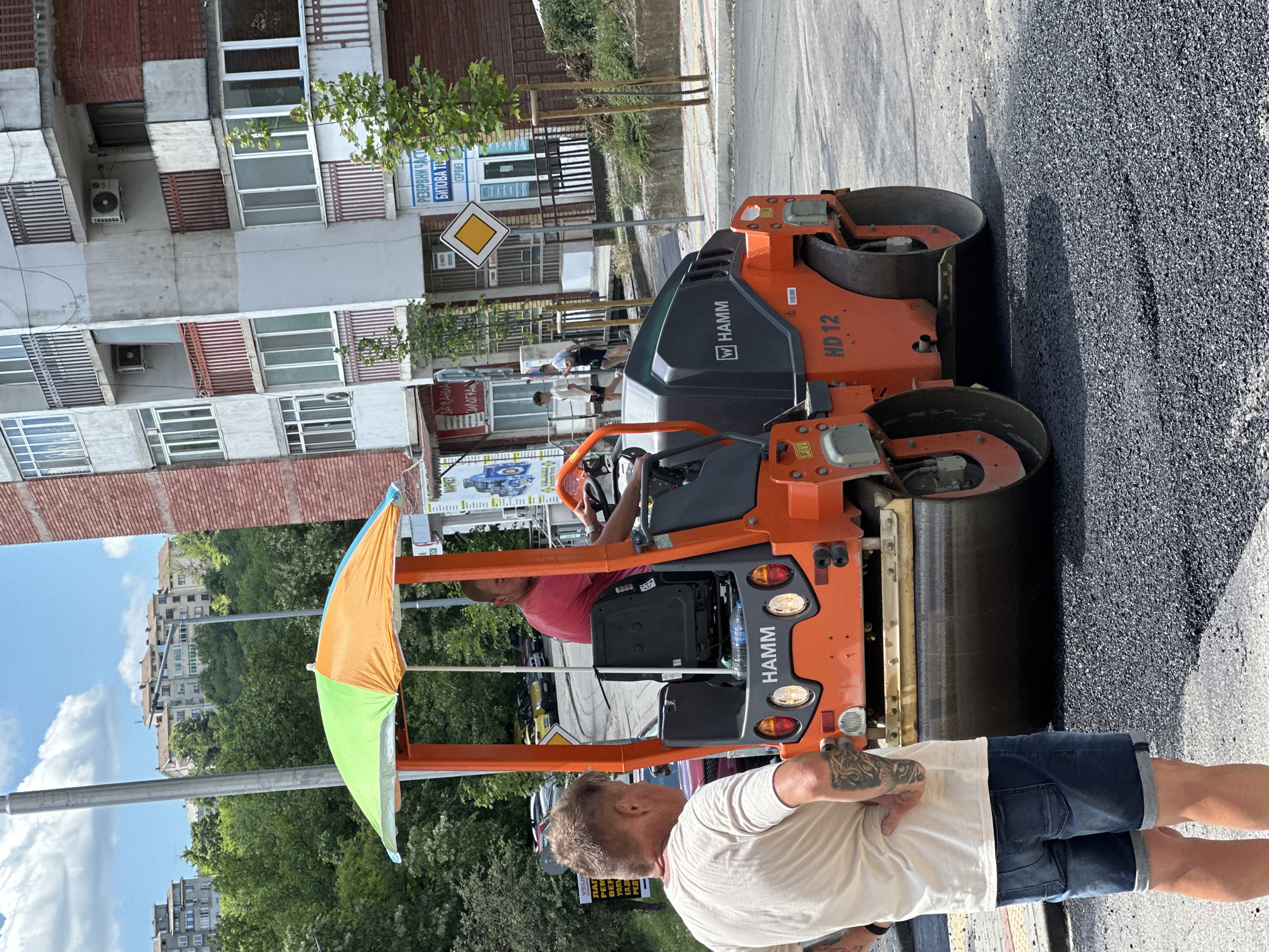 This image depicts a street paving scene in Veliko Tarnovo, Bulgaria. In the foreground, a man with tattoos on his left arm and leg, wearing a light-colored t-shirt and denim shorts, stands with his back to the camera, observing the activity. In the midground, an orange HAMM HD 12 road roller is actively compacting fresh asphalt. A man wearing a red t-shirt is operating the roller. A colorful umbrella, providing shade, is attached to the roller's frame. The background shows a multi-story residential building with balconies and air conditioning units, typical of urban Bulgarian architecture. Green trees and a cloudy blue sky complete the environment. Signs in Cyrillic are visible on the buildings, indicating local businesses or services. The overall scene suggests ongoing municipal work in a residential area of the city.