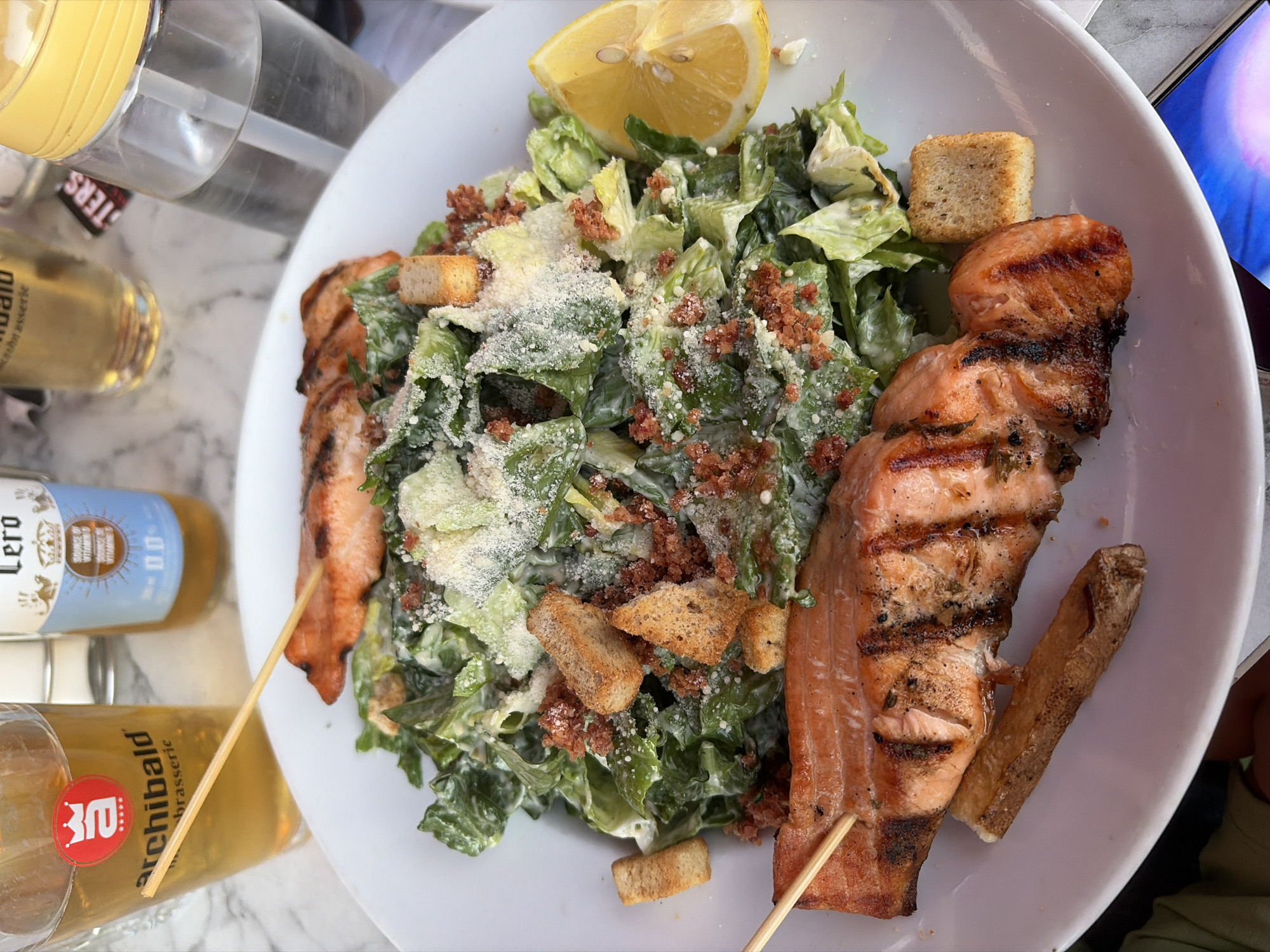 A top-down, close-up shot shows a white plate of food with grilled salmon and salad. Two pieces of grilled salmon are on skewers, one on top of the other, layered over a Caesar salad. The salad contains romaine lettuce, croutons, and shaved Parmesan cheese, with crumbled bacon bits interspersed throughout. A slice of lemon is placed on the side of the salad. In the background, a glass of amber liquid, a bottle of beer with a blue label, and a clear plastic water bottle with a yellow lid are visible on a marble-patterned surface.