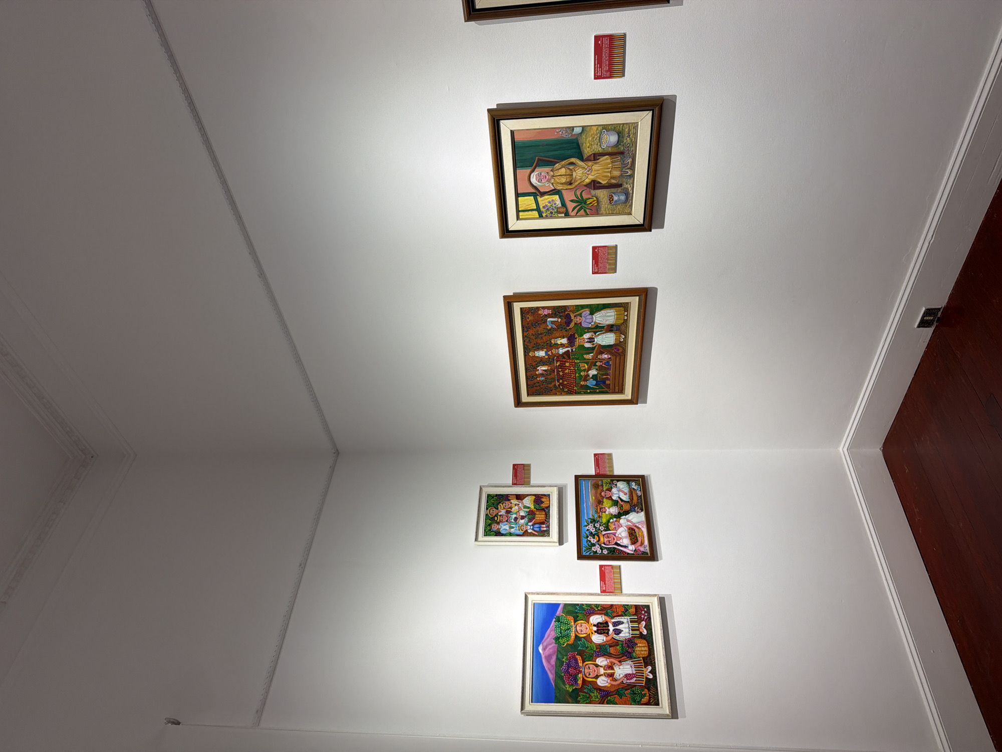 This is a photograph of an art exhibition in La Orotava, Spain, showcasing several paintings displayed on a white wall. The paintings appear to be folk art or naive art, characterized by vibrant colors and depictions of people engaged in daily activities, often with a rural or harvest theme.
On the left wall, there are three framed paintings. The largest one at the bottom left features two women with baskets of fruit on their heads, with a volcano in the background. Above this, a smaller painting shows a group of people carrying baskets. To the right of these, a rectangular painting depicts children and adults with baskets of fruit.
On the adjacent wall, there are two more framed paintings. The one on the left shows a group of people harvesting from trees. The painting on the right is a portrait of an older woman sitting in a chair, wearing a yellow dress and holding a basket. Red informational plaques are visible below some of the paintings.
The exhibition space has white walls and a dark wooden floor. The corner of the room is visible, and there is subtle architectural molding along the ceiling. The lighting appears to be artificial, illuminating the artwork. The overall impression is that of a well-maintained gallery space.