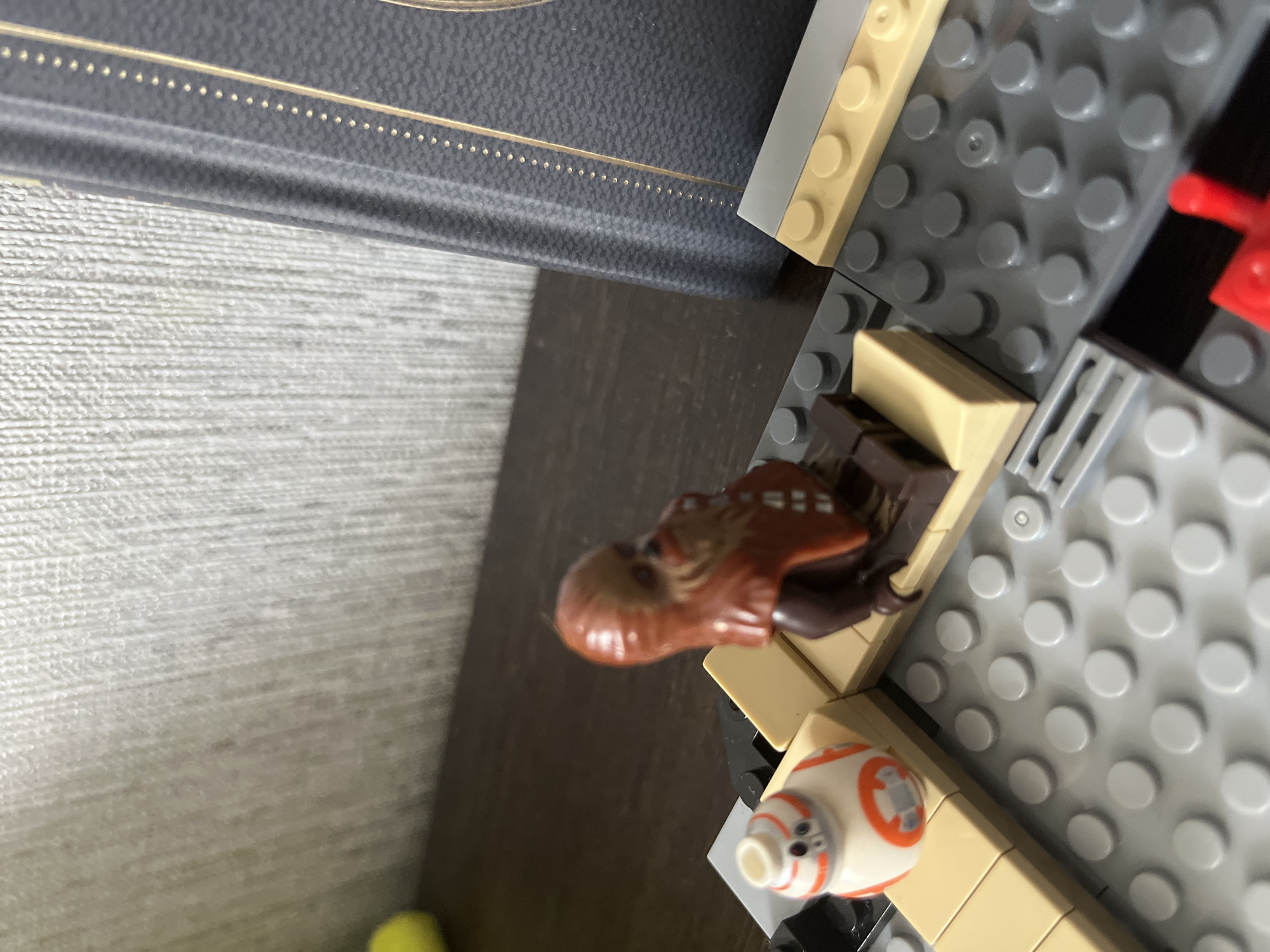 The image shows a close-up view of LEGO Star Wars figurines on a surface that appears to be a shelf or table, with a textured wall in the background and a dark blue book with gold trim to the right. In the foreground, a LEGO BB-8 droid is visible on a beige LEGO brick structure. Adjacent to the BB-8 is a LEGO Chewbacca figurine seated on a brown LEGO brick piece. The LEGO bricks are arranged in a grey baseplate. No information about the city of Saratov, Russia is present in the image.