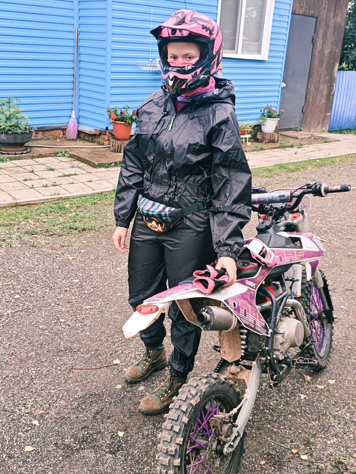 A young female with auburn hair is standing next to a dirt bike. She is wearing a black rain suit with a pink and black helmet. A fanny pack with a colorful pattern is around her waist. Her boots are brown and green. The dirt bike is predominantly pink and white with purple accents on the wheels. The background consists of a blue building, a fence, and some potted plants. The ground is covered in gravel. This scene is likely located in Rassol'naya, Russia, given the context provided.