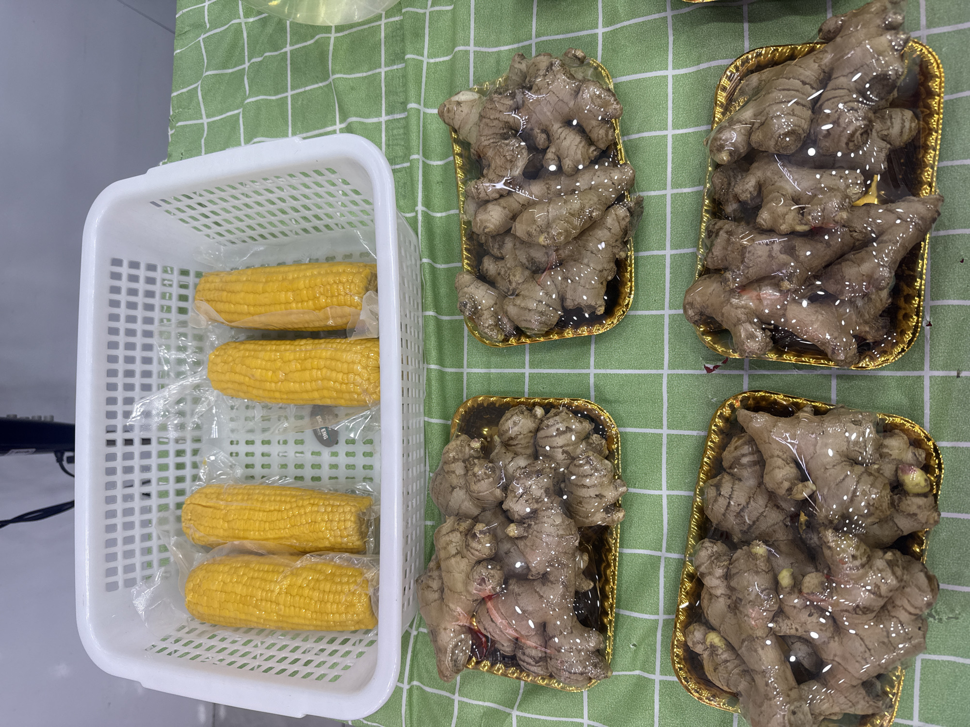 The image depicts a countertop covered with a green and white checkered tablecloth. A white plastic basket containing four ears of corn wrapped in plastic is positioned to the left. To the right of the basket are four gold-colored trays, each containing a mass of ginger roots also wrapped in plastic. The context for the location in the city of Yuzhong District, China is not evident from the image itself.