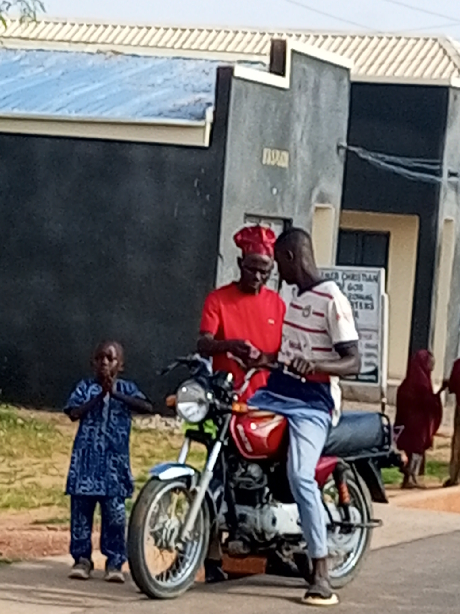 In Gombe, Nigeria, a street scene unfolds with four individuals and a red motorcycle positioned on a paved surface adjacent to a building. The building has a dark gray facade and a lighter gray section, with a cream-colored trim along the roofline. A sign is affixed to the lighter gray section of the building.
In the foreground, a red motorcycle is parked, with its front wheel and part of the engine visible. A young boy, dressed in a blue patterned tunic and trousers, stands to the left of the motorcycle, with his hands clasped together.
To the right of the boy and slightly behind him, two men are engaged in conversation. The man on the left wears a red tunic and a red cap. The man on the right is in a white and red striped t-shirt and blue trousers. Both men appear to be looking at something held in their hands, possibly money.
Further to the right and in the background, a person wearing a dark red headscarf and a dark robe is visible, partially obscured. Another figure, mostly out of frame, is also present in the background. The ground is covered with a mixture of dirt and grass, suggesting an outdoor setting.