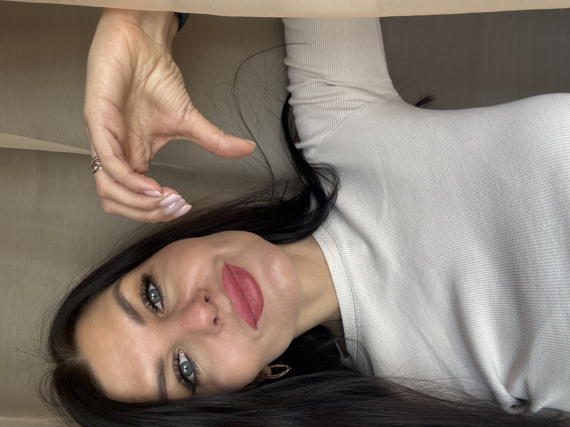 The image is an indoor selfie taken from a rotated perspective. A woman with fair skin and dark hair is facing the camera. Her eyes are light-colored with dark mascara. She has defined eyebrows and a nose piercing. Her lips are a matte red color. She is wearing a light-colored long-sleeved shirt. Her left hand is raised near the top left of the frame, displaying a ring on one finger and a manicure with light pink nail polish. The background appears to be a beige-colored surface. There is no other information in the image to confirm the location is Perm, Russia.
