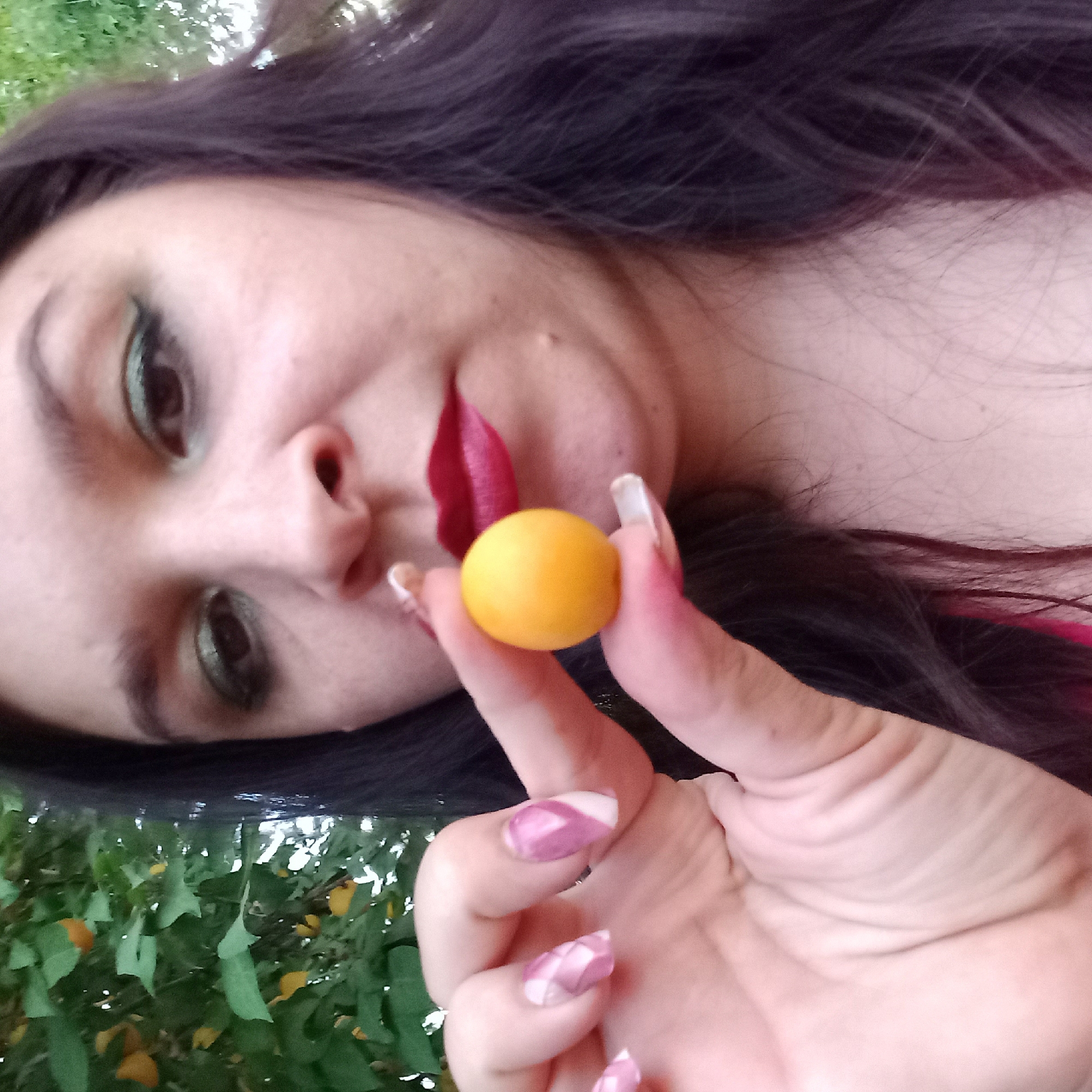 A woman, possibly in Berezhynka, Ukraine, holds a small, round, yellow fruit between her thumb and forefinger. She is wearing red lipstick and green eyeshadow. The background consists of green foliage. Her fingernails are painted with a pink and white design.