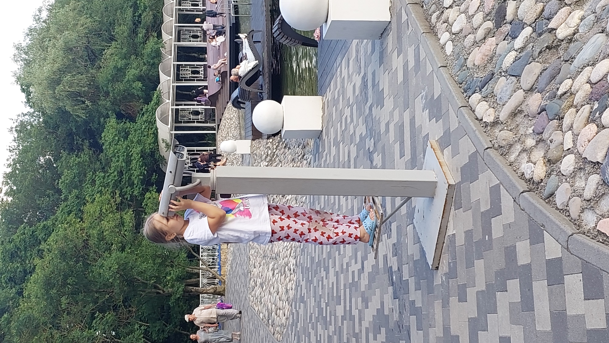In a park setting, a young girl with blonde hair is looking through a telescope, positioned on a paved walkway. She is wearing a white t-shirt with a colorful graphic and pink pants adorned with red butterfly patterns. Her bright blue sandals are visible. The walkway is made of grey and dark grey interlocking pavers, bordered by a gravel area filled with various colored stones.
Behind the girl, a body of water reflects the overcast sky, suggesting it might be a cloudy day. In the background, there is a wooden platform with people relaxing on benches, some appearing to be enjoying the view. A white gazebo-like structure with decorative railings is also present, partially obscured by trees. Further in the distance, more trees create a lush green backdrop.
The scene suggests a leisurely day outdoors. The girl's focused expression indicates her engagement with the telescope, perhaps exploring the surroundings or observing something of interest. The overall atmosphere is peaceful and recreational. There is no visible text in the image.