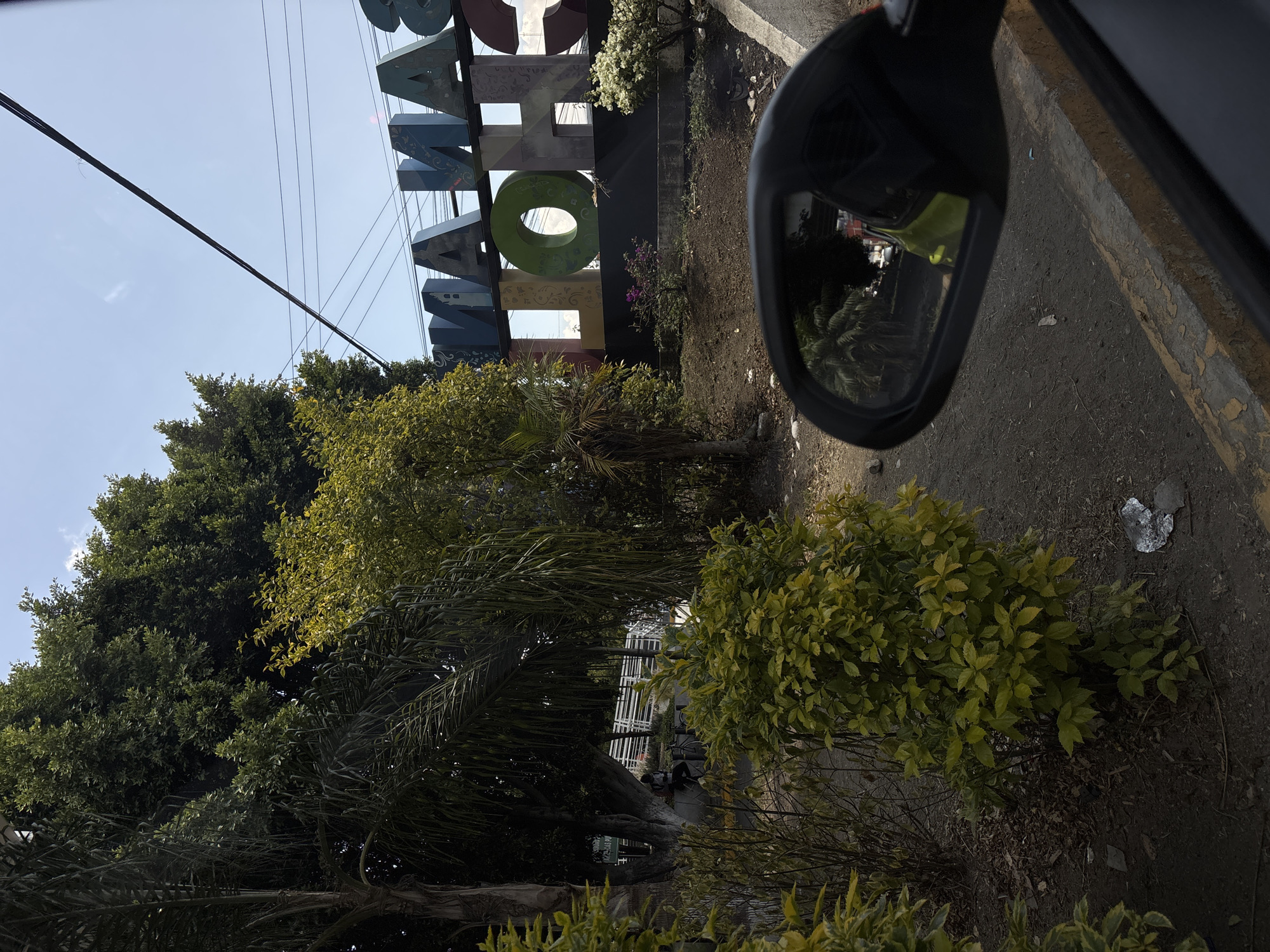 The image shows a view from inside a car, looking out towards a landscaped area in San Andrés Cholula, Mexico. A large, colorful sign spelling out "SAN ANDRÉS CHOLULA" is visible in the background. The sign is made of individual block letters in various colors and decorative patterns. In the foreground, there are various trees and bushes. A car side mirror is in the right side of the frame. The street is on the right and there is a sidewalk there. Power lines run across the sky.