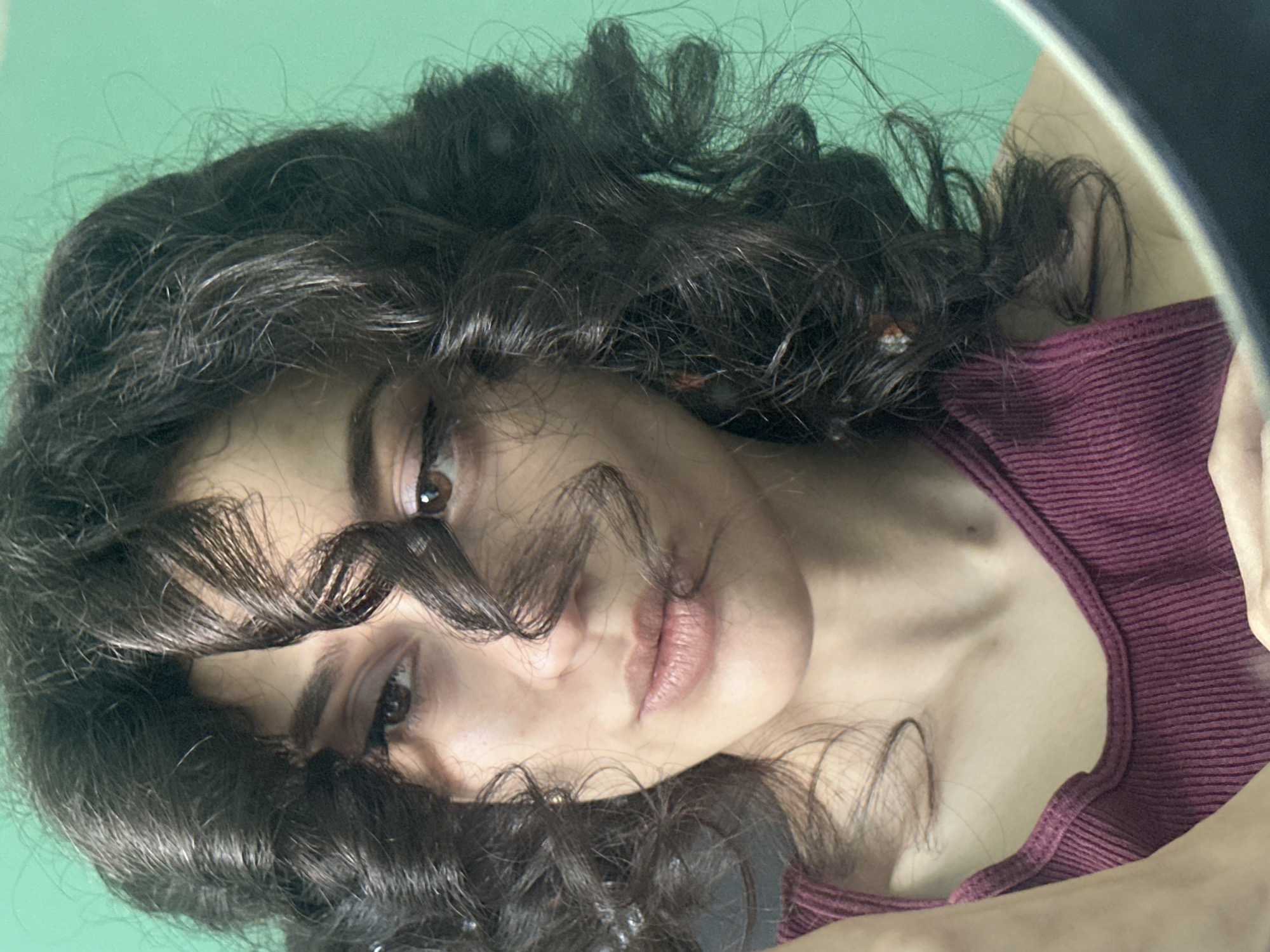 A close-up, waist-up portrait of a young woman with dark, curly hair framing her face is shown against a bright green background. She is wearing a maroon ribbed tank top. Her gaze is directed slightly to her left, with a subtle smile. Her left hand is visible in the lower left corner, holding a mirror, indicating the photo was taken in front of one. The lighting suggests an indoor setting, potentially a home, with the green background being a wall or backdrop. There are no discernible landmarks to pinpoint a specific location within Sofia, Bulgaria.