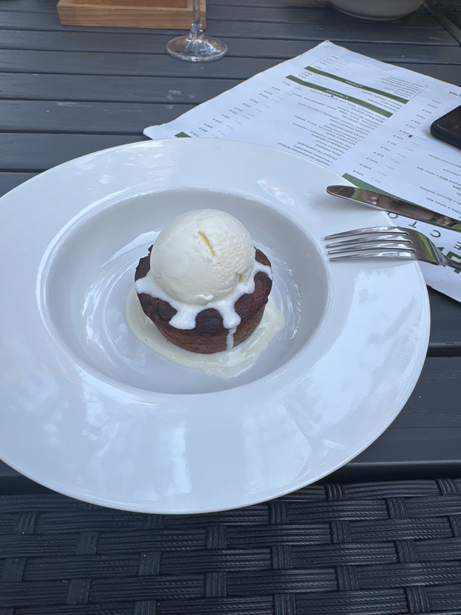 A close-up shot features a decadent dessert, likely a chocolate lava cake with a scoop of vanilla ice cream, artfully presented in a large white ceramic bowl. The cake appears moist with melted chocolate oozing around it, and the ice cream is melting slightly, creating a creamy sauce. The dish is set on a dark, textured surface, possibly a table made of woven material, characteristic of outdoor seating at a restaurant. In the background, a portion of a menu is visible, with text in Russian, suggesting the location is in Russia. A fork and knife rest on the menu, indicating the dessert is ready to be enjoyed. The overall lighting suggests it could be daytime, possibly outdoors or in a well-lit indoor space. Without further context or visual cues of specific landmarks, pinpointing the exact location in Novosibirsk, Russia, is not possible from this image alone.