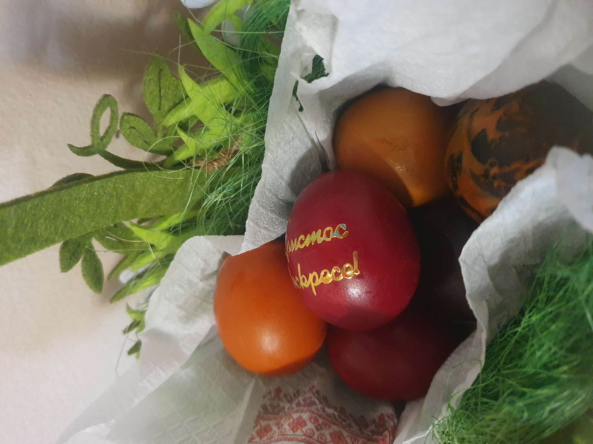 The image depicts a collection of Easter eggs, likely in Plovdiv, Bulgaria, given the Cyrillic inscription "Христос воскресе!" ("Christ is risen!") on one of the red eggs. The eggs are primarily orange and red, with one egg having a speckled, brown and orange pattern. They are nestled inside a white, crinkled paper container or bag. The arrangement also includes decorative elements such as green, grassy-looking filler and felt-like green leaves. A portion of a red and white patterned textile is visible at the bottom of the container.