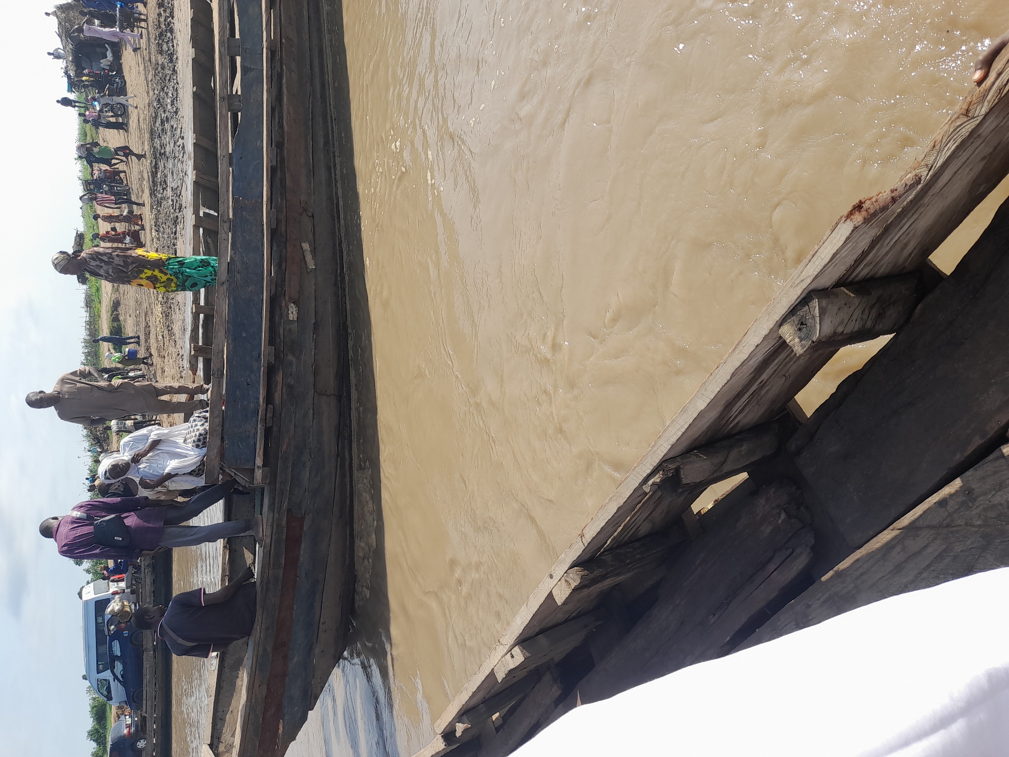 In Ibi, Nigeria, a bustling scene unfolds at a riverbank, where several wooden boats are moored. The murky, brown water churns with visible ripples, suggesting a current. In the foreground, the edge of another wooden boat is visible, its weathered planks showing signs of age and use.
On one of the boats, a group of people are gathered. A man in a purple shirt and grey trousers stands, facing away from the camera, with a dark bag slung over his shoulder. Next to him, a man in a dark shirt and red-trimmed collar is seated, looking to his left. Further back on the same boat, a woman in a white head covering and patterned skirt is seated, looking towards the left. Another man, dressed in a light brown garment, stands near the edge of the boat.
On the sandy bank behind the boats, more people are visible. A woman in a colorful, patterned dress stands prominently, facing left. Further in the background, there are several individuals, motorcycles, and a makeshift shelter with a thatched roof. A white van and a dark blue car are parked on the left side of the image, indicating vehicular access to the area. The sky above is partly cloudy. The overall impression is one of a lively, functional riverside community engaged in daily activities.