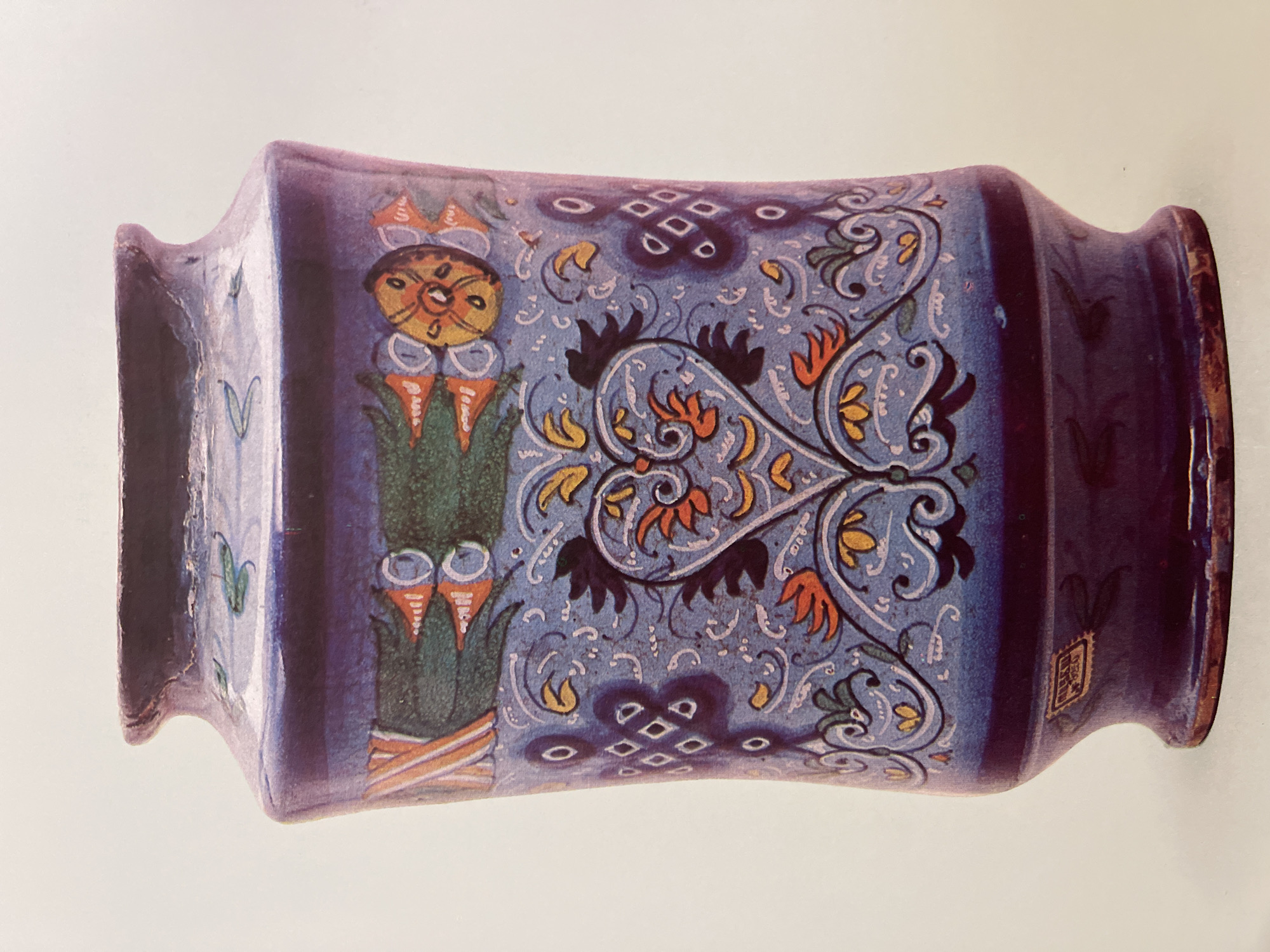 The image shows a ceramic vase with a rectangular body and flared rims. The vase is primarily blue with intricate decorative patterns in various colors including yellow, orange, green, and black. The patterns include floral motifs, geometric shapes resembling knots, and stylized plant designs. A small, square emblem is present on the lower right side. The vase is photographed against a light background. This artifact can be found in the A. Kasteyev State Museum of Arts in Almaty, Kazakhstan.