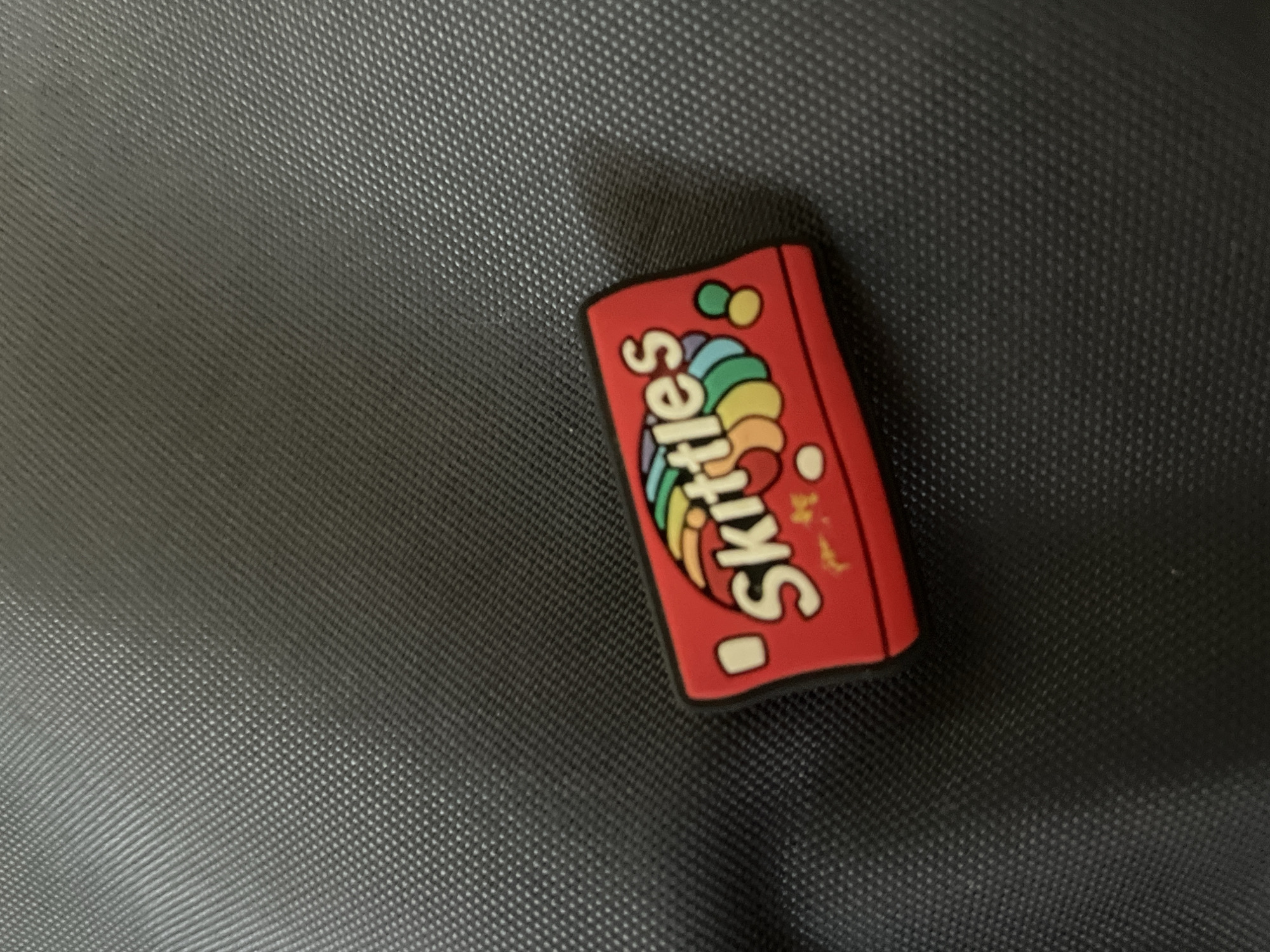 A close-up, still-life image captures a small, red, rectangular object resembling a miniature Skittles candy package, resting on a dark, textured surface. The object, likely a charm or decorative piece made of rubber or silicone, features the brand name "Skittles" in white lettering prominently displayed on its front. A colorful rainbow graphic is visible underneath and behind the text, accompanied by scattered small, round shapes in green, yellow, light blue, and white, consistent with the candy's branding. Faint yellowish marks are visible on the right side of the red object.
The background is a dark, possibly black or very dark grey, surface with a distinct, repeating grid-like pattern of small raised dots, creating a tactile texture. The lighting is soft and even, casting subtle shadows to the left and bottom of the object. There are no people or discernible activities in the frame, and the setting appears to be an indoor or controlled environment. The specific location of Baranovka, Russia, is provided as context but cannot be visually verified from the image itself.