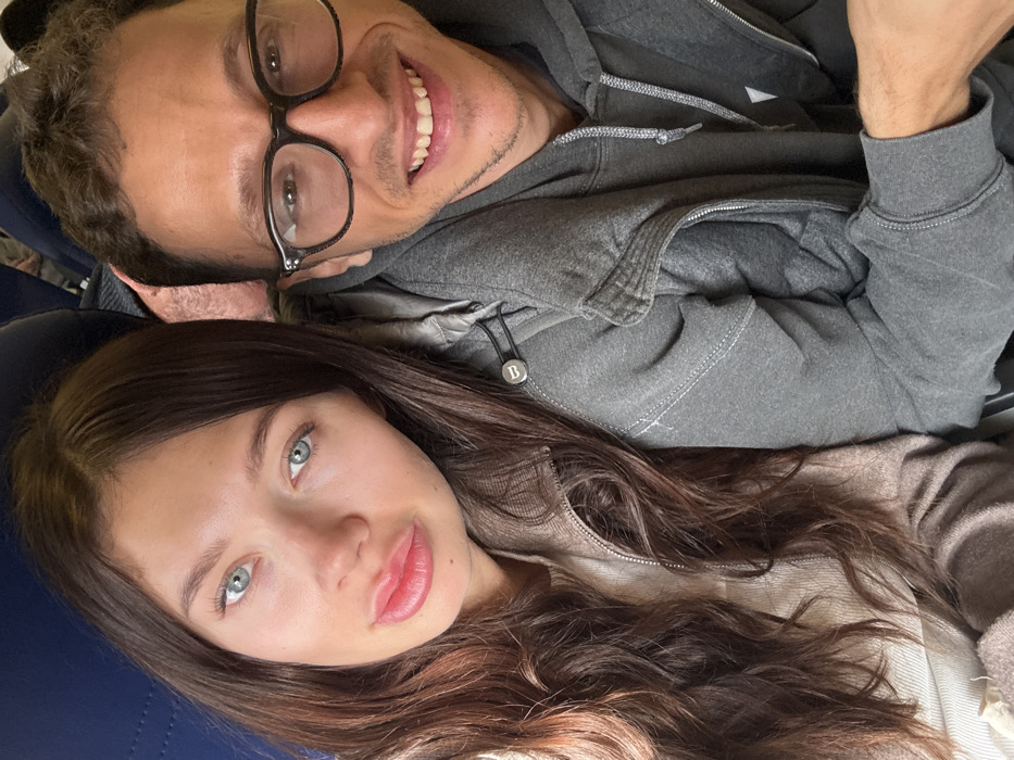 The image displays two individuals, an adult female and an adult male, seated in close proximity within what appears to be the cabin of a passenger aircraft, indicated by the blue upholstered seating visible in the background. The location is Colombier-Saugnieu, France.
The female, positioned on the left, has long, wavy dark brown hair, light blue-green eyes, and full lips, which are slightly parted. She is wearing a light beige or off-white ribbed top beneath a light brown or taupe zippered jacket, which features a circular dark button with a white 'B' emblem on the shoulder area. She looks directly towards the camera with a neutral expression.
The male, situated to the right and slightly behind the female, has short, dark, curly hair and is wearing dark-framed eyeglasses. He smiles broadly, revealing his teeth, and is dressed in a grey hooded sweatshirt with visible white stitching details on the seams. Both individuals are looking towards the camera. No specific actions beyond posing for the photograph are depicted.