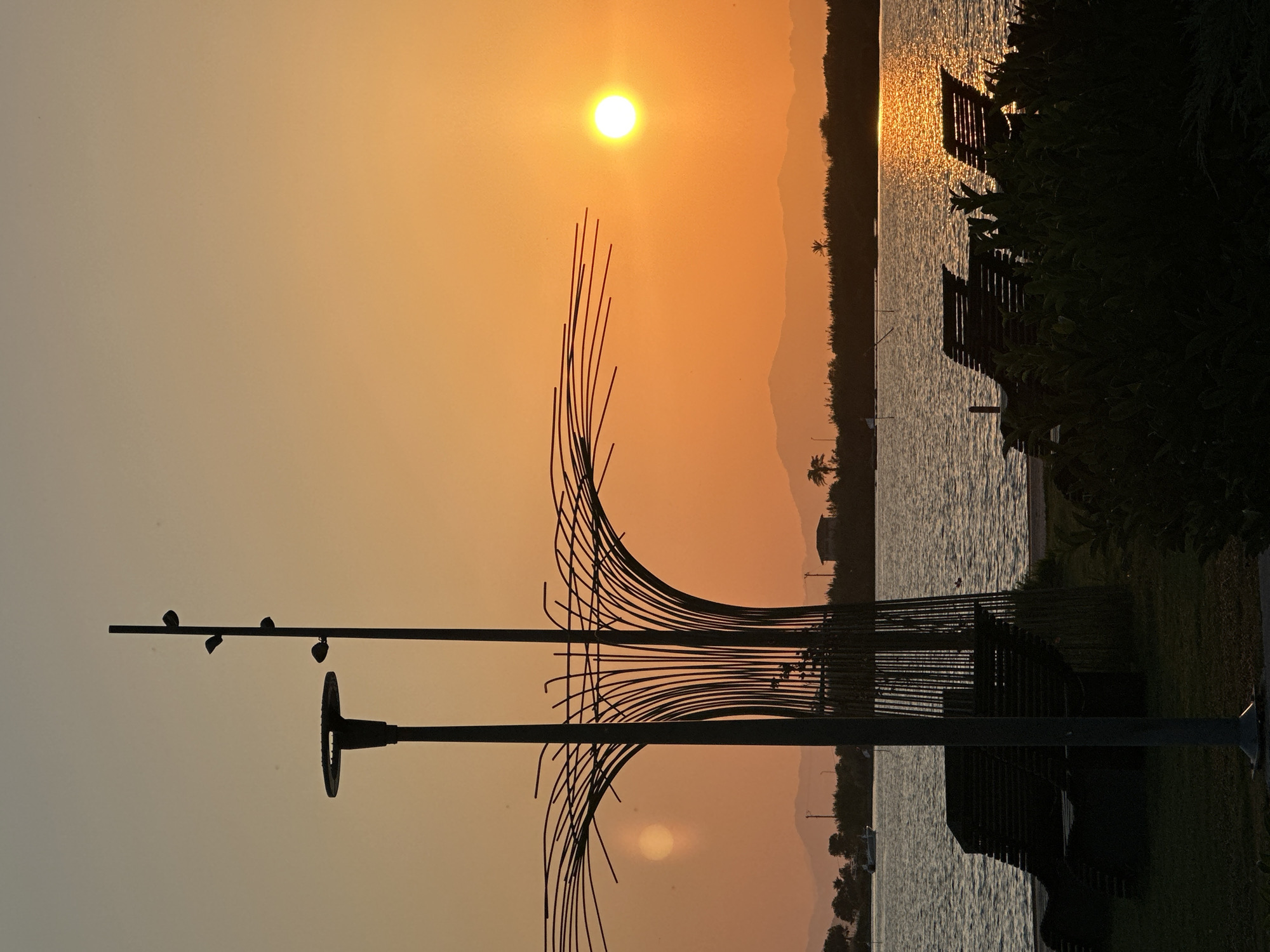 The image depicts a sunset over a body of water, likely a bay or harbor, in Fethiye, Turkey. The sky is a warm gradient of orange and yellow, with the sun visible as a bright, luminous orb. In the foreground, a silhouetted sculpture made of thin, curving metal rods dominates the scene. To the right of the sculpture, there are dark green trees and what appears to be a park bench or seating area, also in silhouette.
The water is rippling, reflecting the golden light of the setting sun. In the distance, a dark, tree-lined horizon is visible, with faint outlines of hills or mountains behind it. There are also silhouetted palm trees along the shoreline, indicating a warm climate. A lamppost with several lights is also visible in silhouette, along with a sculptural element that resembles a large, stylized flower or fan. The overall mood is peaceful and serene, capturing the beauty of a sunset in a coastal setting. The time of day is clearly sunset due to the position of the sun and the warm, golden light. The weather appears to be clear and calm. There is no visible text in the image.