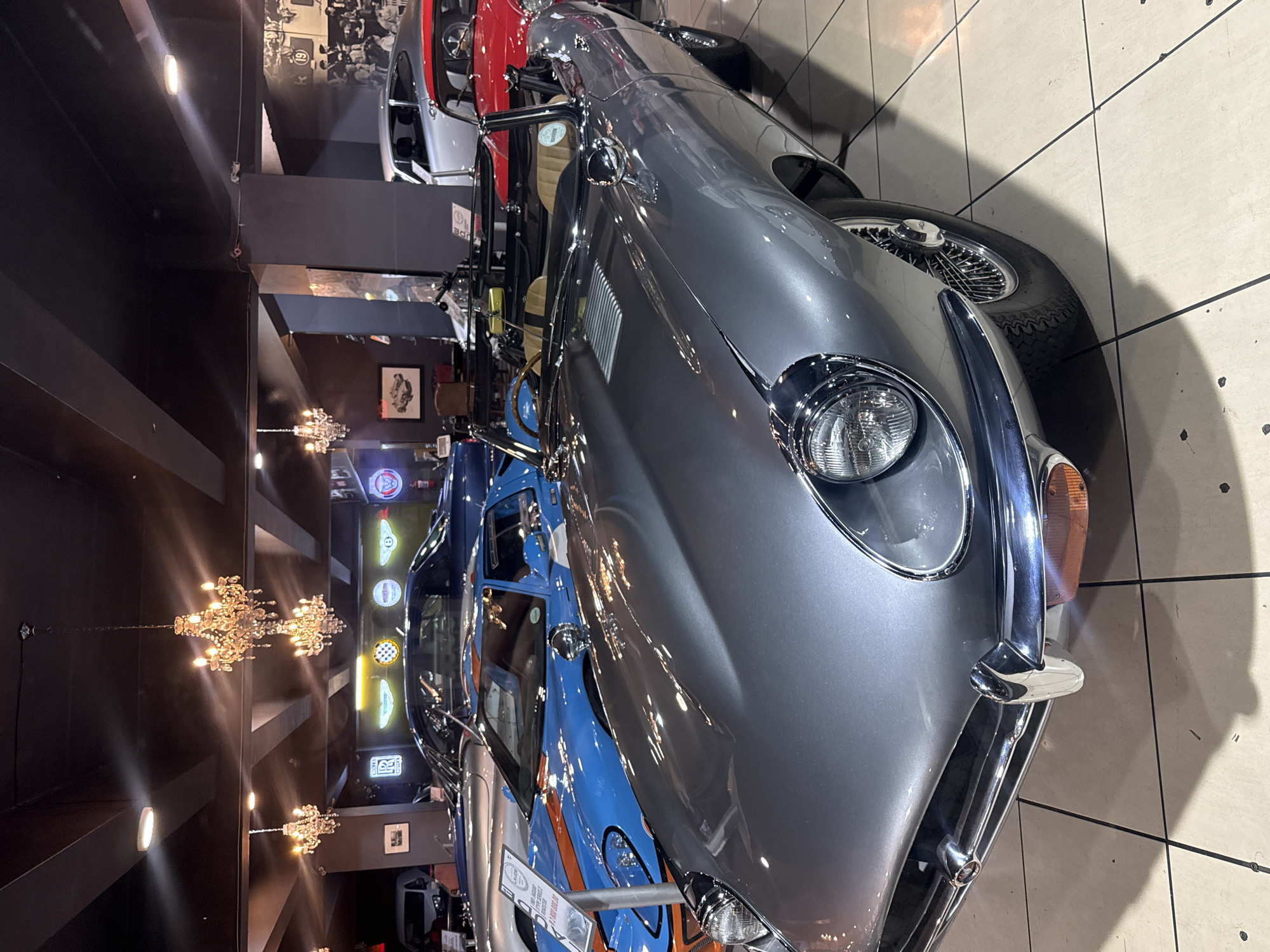 A photograph shows a gray vintage car in an indoor showroom, possibly a museum or private collection, in Cape Town, South Africa. The car, a classic model with wire wheels and chrome accents, is parked on a tiled floor. In the background, there are more cars stacked vertically and illuminated signs, along with chandeliers hanging from the ceiling. The walls are dark, suggesting the location is enclosed and designed to showcase the vehicles.