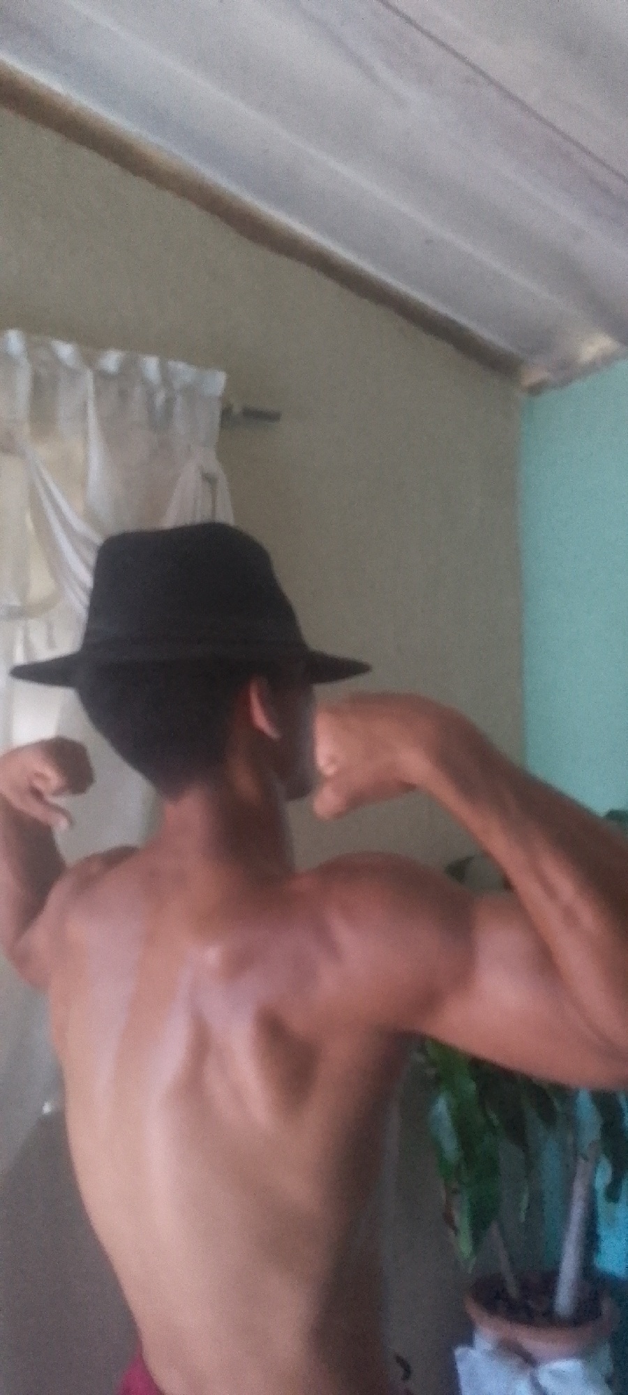 A shirtless, muscular man wearing a black fedora hat is flexing his biceps and back muscles. He is standing indoors, possibly in a home. In the background are white curtains, a wall painted in light colors (possibly beige and light blue), and a potted plant. I have no information that supports this picture was taken in the city of Juan Griego, Venezuela.