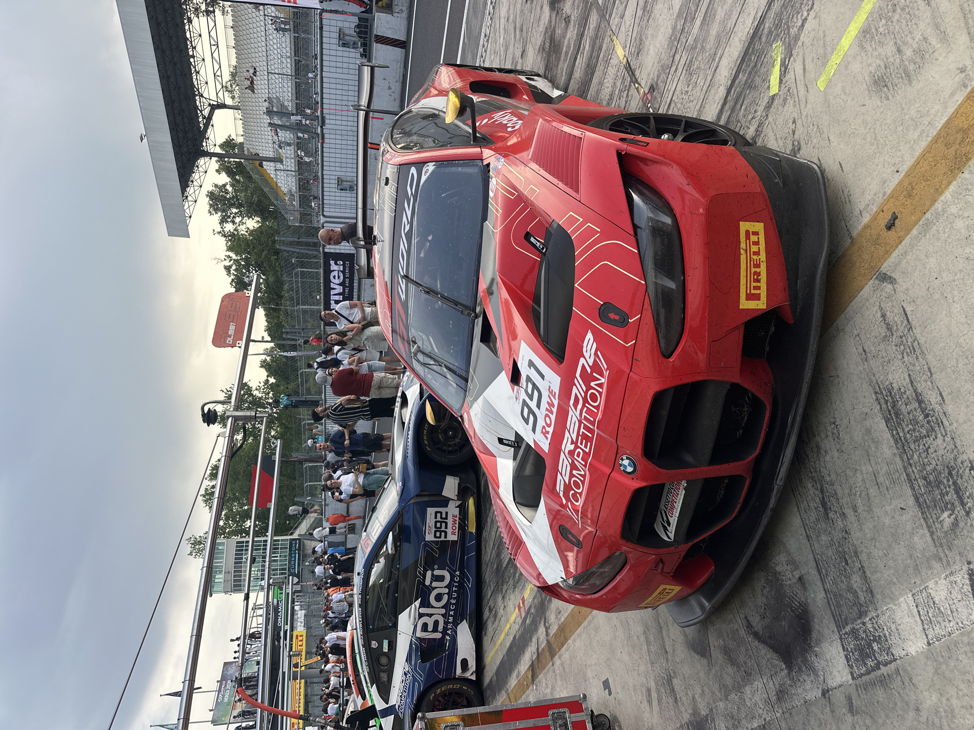 A race car, numbered 991, is parked next to another race car, numbered 992. The race cars are stationed in front of a crowd of people. The cars are labeled "ROWD PARADINE COMPETITION," "ASSETTO CO COMPETIZIONE" and "Blau FARMACÊUTICA," and "d river TYRE AND SERVICE." The background shows an observation tower and the sky. There is a yellow line on the ground. The location is Monza, Italy.