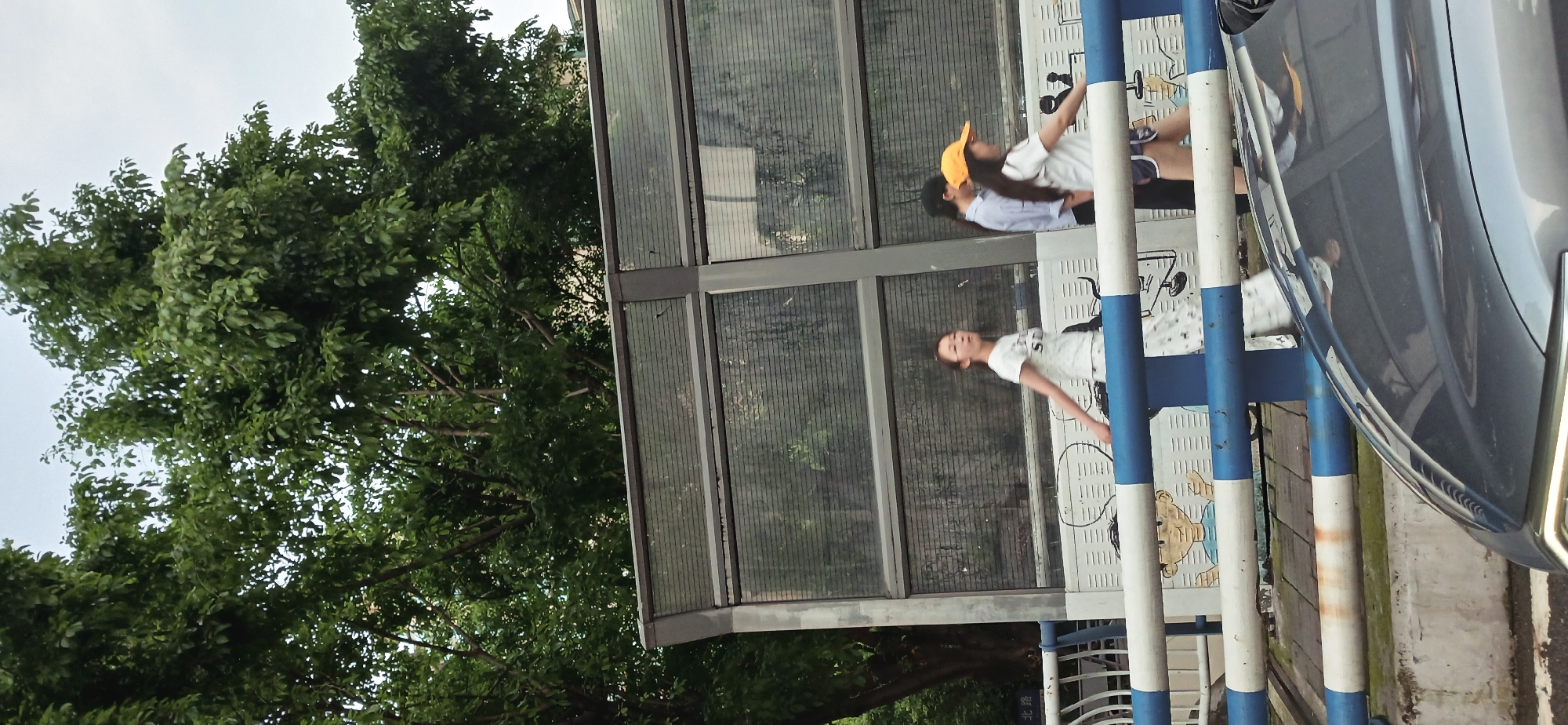 The image depicts an outdoor scene in Yuzhong District, China. A bus stop structure is visible, with a woman wearing a white t-shirt and patterned pants standing near the stop. Behind her, partially obscured, are two more people, one wearing a yellow hat. The bus stop's design features blue and white striped poles and a patterned surface. A vehicle, possibly a car, is parked nearby, its reflection visible on the bus stop. The environment includes a tree with dense green foliage.