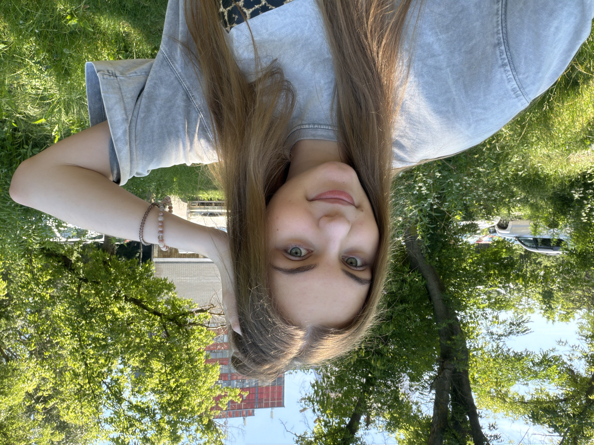 This is a close-up, upside-down selfie of a young woman with long, light brown hair and green eyes. She is wearing a light blue, oversized t-shirt and a bracelet on her left wrist. The background shows a sunny outdoor setting with green grass, trees, and buildings. The woman appears to be smiling or smirking faintly, with a relaxed expression. The lighting suggests it is daytime, likely a clear, sunny day. In the distance, there are some buildings, including one with red accents, and a vehicle can be seen. The overall impression is of a casual, outdoor snapshot taken in an urban or suburban environment.