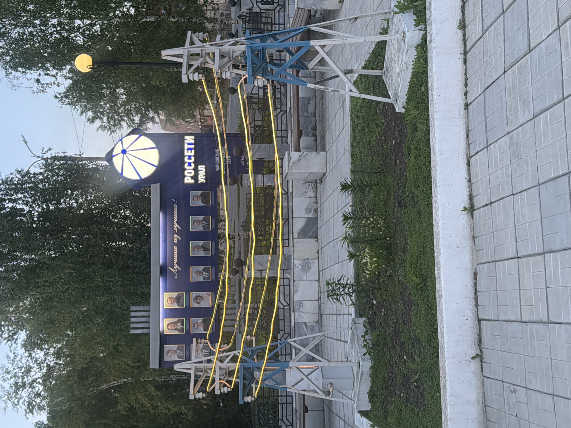 The image shows a monument or display in the city of Chaikovsky, Russia. The main feature is a blue sign with the words "РОССЕТИ УРАЛ" (Rosseti Ural) and "Лучшие из лучших!" (Best of the best!). Below the text are rows of portraits, likely recognizing individuals associated with the energy company Rosseti Ural.
To the left of the sign is a street lamp, and in front of the sign are decorative replicas of power line towers painted white and blue. The structures are connected by several yellow lines that appear to be strings of lights.
The monument is situated on a paved area with green patches of grass and small plants along the edge. The overall setting suggests a public space, possibly a park or a town square. The angle of the image suggests it was taken from a nearby street.