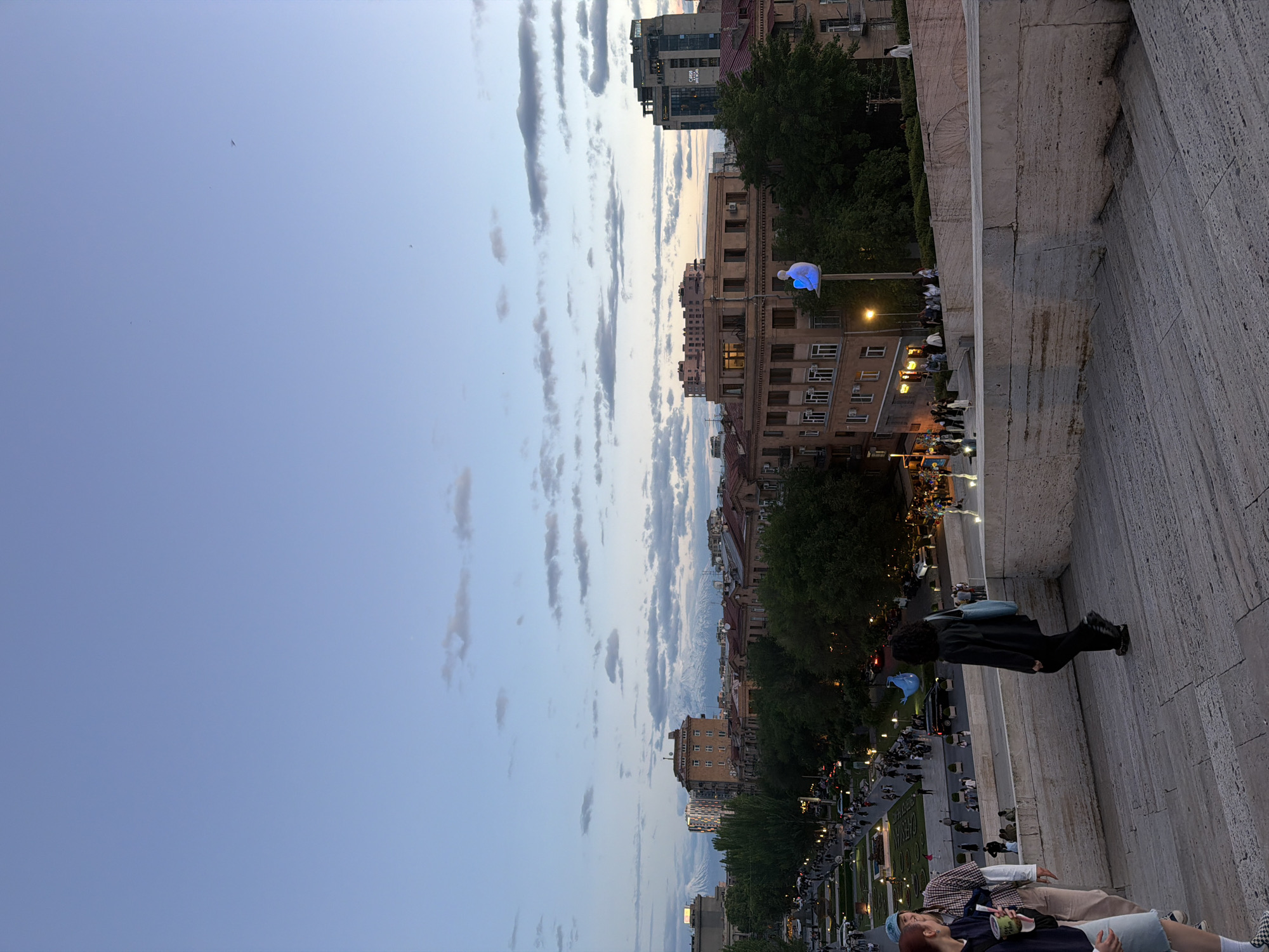 The image depicts a view in Yerevan, Armenia, likely captured at the Cascade complex. The scene presents a descending perspective from an elevated section of the Cascade's stairway, which is constructed of light-colored stone and exhibits visible wear and tear.
Several people are present, some sitting on the steps and others walking below on what appears to be a lower level of the Cascade or a surrounding park area. At least one person is reclining on the stairs, and another is standing and looking toward the street below.
The cityscape is visible beyond the Cascade. Buildings of varying heights and architectural styles are present, and green trees line the street. The sky is partly cloudy. The image appears to have been taken during twilight, judging by the ambient light levels. A light fixture or decorative element glowing blue is visible among the trees and buildings.