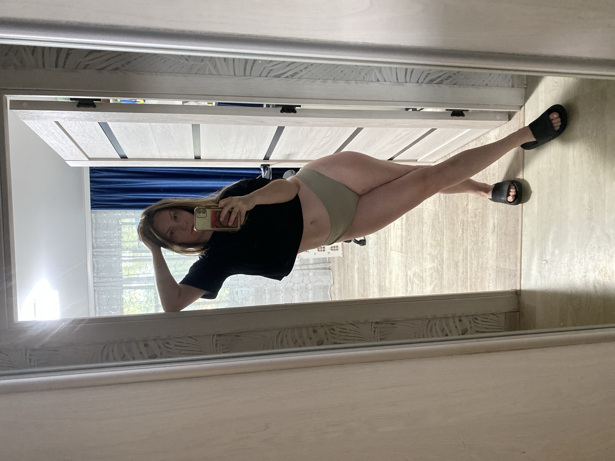 A woman is taking a selfie in a mirror. She is wearing a black shirt and olive-colored underwear. She is also wearing black slides on her feet. The background shows a doorway with a white door, a blue curtain, and a window. The mirror frame is also white. The location is likely indoors in Noviy, Russia.