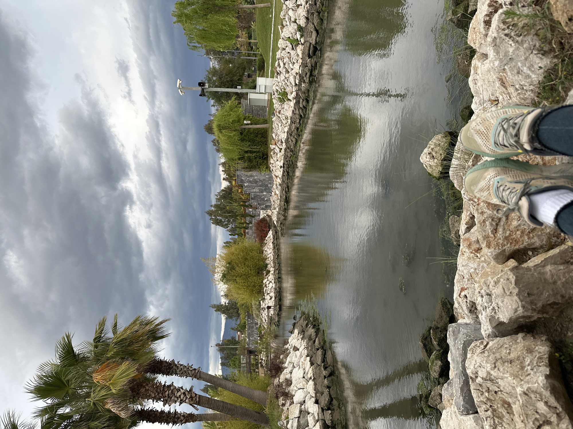 A long shot shows a body of water, possibly a canal or narrow pond, with stone banks. A pair of shoes, worn by an unseen person, are visible on the right bank. In the background, the sky is heavily clouded with dark, gray clouds. The vegetation along the banks includes trees, palms, and shrubs. A palm tree is prominently positioned in the lower left corner.
The location is likely the Fethiye Municipality Park in Fethiye, Turkey. It's a popular park and has a small canal or waterway.