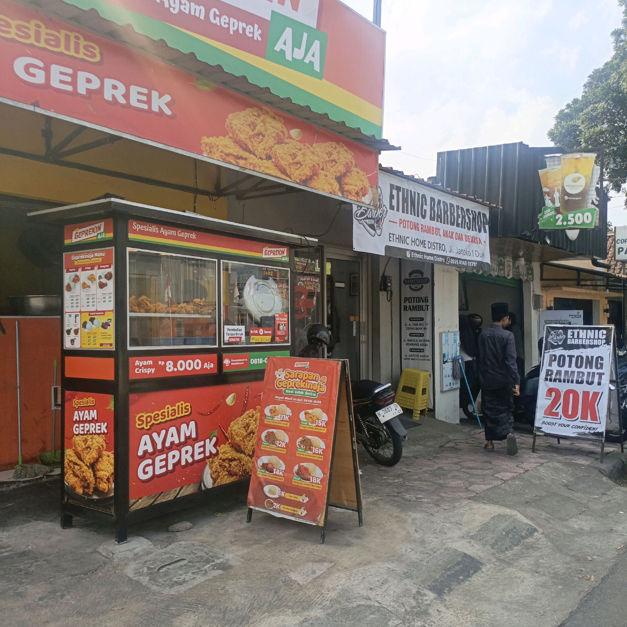 In Salatiga, Indonesia, an outdoor scene captures a food stall and a barbershop. A red and yellow food stall, identified by "GEPREKIN AJA" and "Spesialis Ayam Geprek," displays fried chicken and a menu with prices ranging from 8,000 to 18,000 Rupiah for various chicken dishes. Adjacent to the food stall, a barbershop, "ETHNIC BARBERSHOP," advertises hair cutting services for men, women, and children, with a prominent sign indicating prices starting at 20,000 Rupiah. A person wearing traditional Islamic attire, including a black peci cap and sarong, is seen near the barbershop entrance. Another person, partially visible, is also present. The environment is an urban street with a concrete pavement, some trees in the background, and parked motorcycles. The sky is visible, suggesting it is daytime.