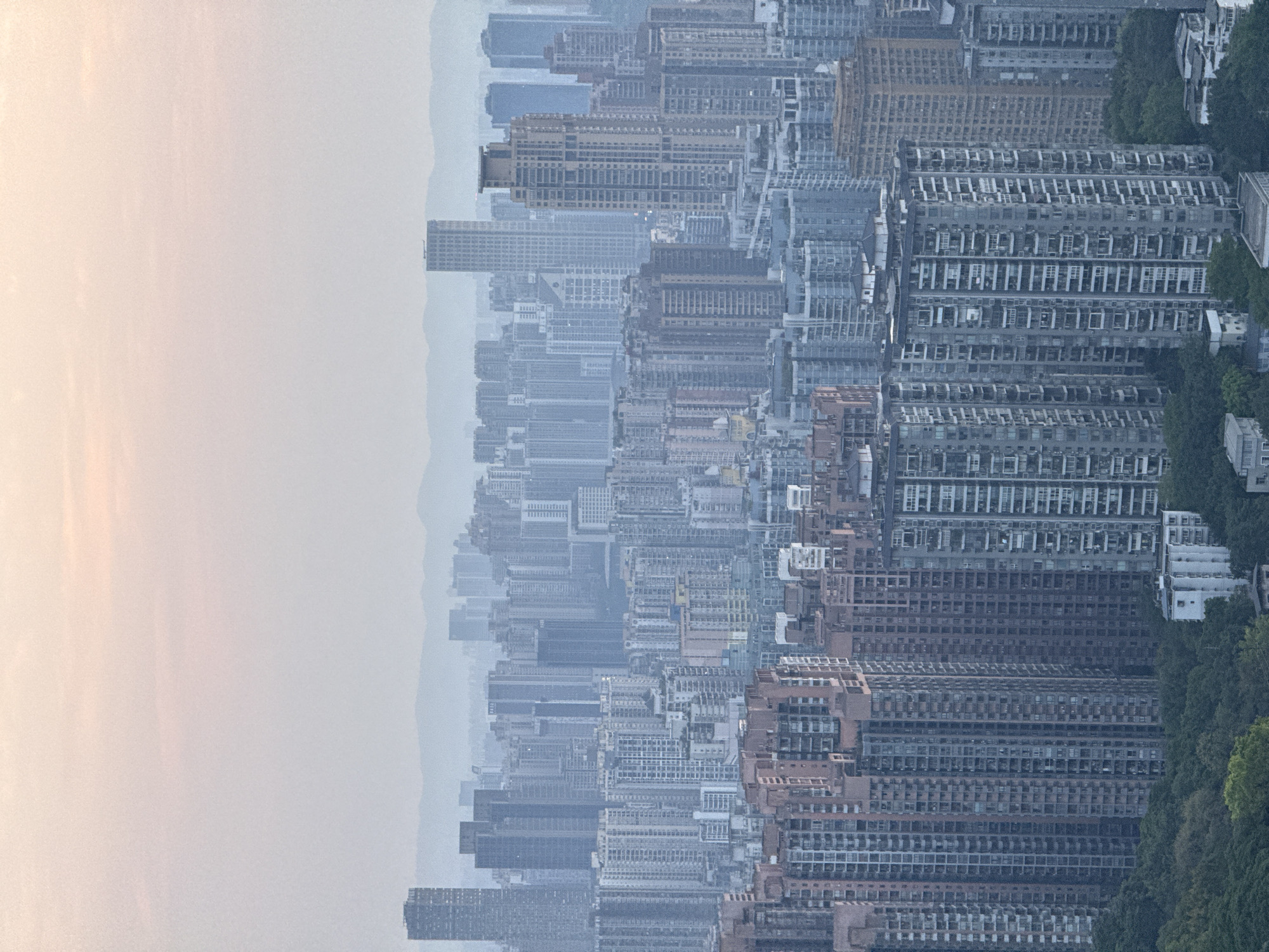 The image presents a dense urban cityscape, dominated by numerous high-rise residential buildings. The structures appear to be tiered and densely packed together, suggesting a high population density. Haze or smog obscures distant buildings on the left. A verdant area with trees is on the right, suggesting this area is on the edge of the city. The image is rotated 90 degrees clockwise.